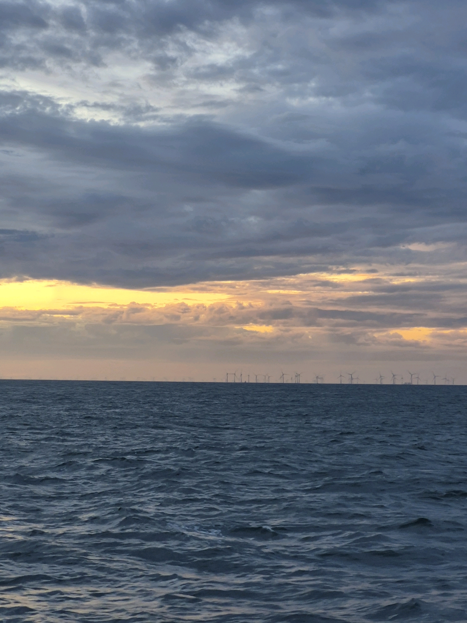 This image depicts a vast expanse of dark blue, choppy water under a cloudy sky. A band of bright yellow and orange light is visible on the horizon, suggesting either sunrise or sunset. In the distance, a line of wind turbines stands silhouetted against the sky, indicating an offshore wind farm. The location is identified as Unknown, Germany, though no specific landmarks or urban features are present to confirm this. There are no people or animals visible in the frame. The overall atmosphere is somewhat dramatic due to the moody sky and the stark industrial presence of the wind turbines.