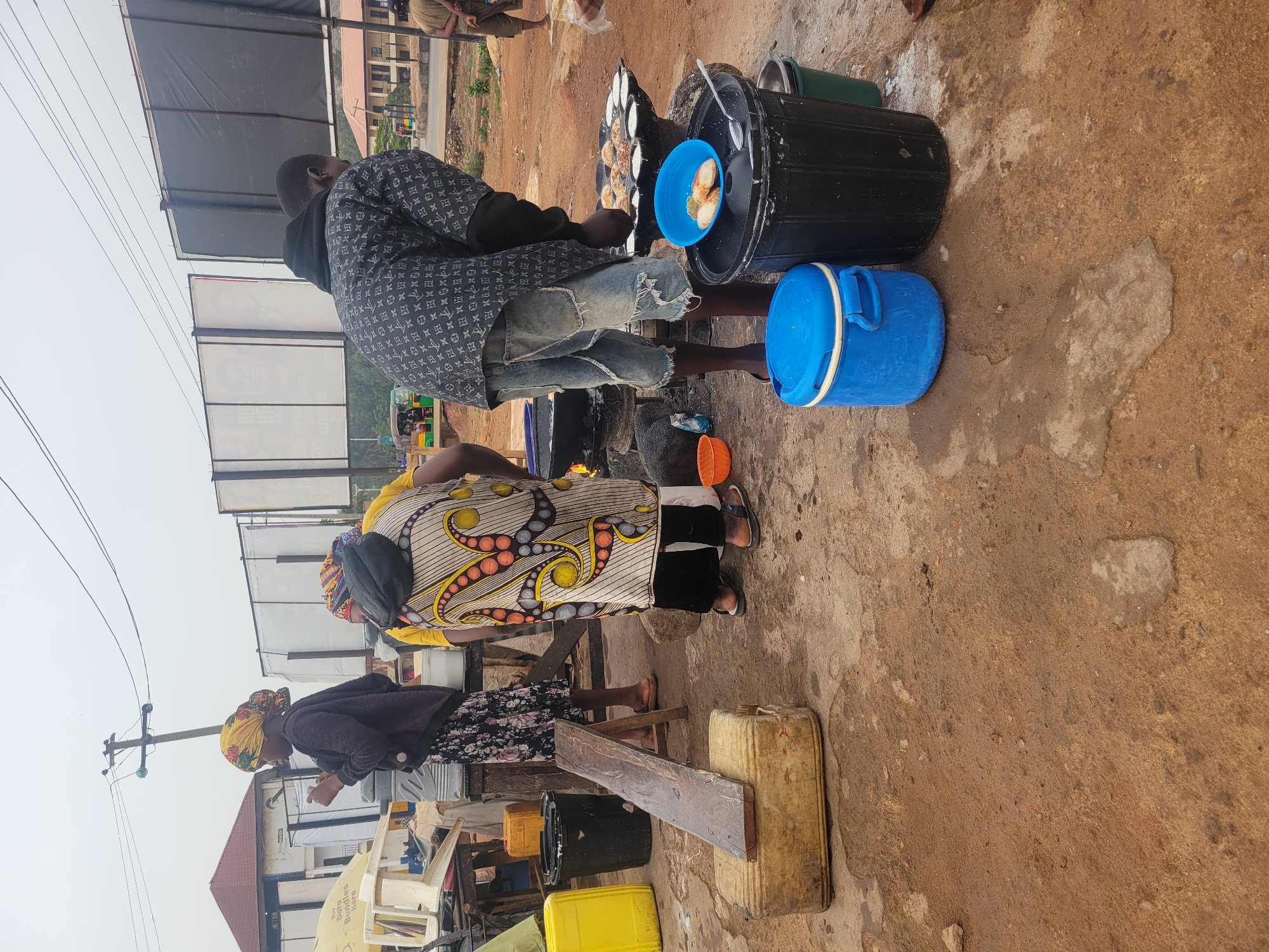 In an outdoor location in the city of Unknown, Unknown, three individuals are present. One individual, a man, is positioned at a cooking setup consisting of a portable stove, a blue bowl with food, and several containers. He is wearing a patterned shirt and ripped jeans, actively engaged in cooking. Adjacent to him is a woman dressed in a patterned dress and black pants, standing as if observing the cooking process. A second woman, with a head covering and dark clothing, stands at a separate makeshift table constructed from containers and a wooden plank, possibly eating or engaged in a transaction. The environment appears to be a street-side setting with nearby buildings and utility poles visible. The ground is dirt and concrete.
