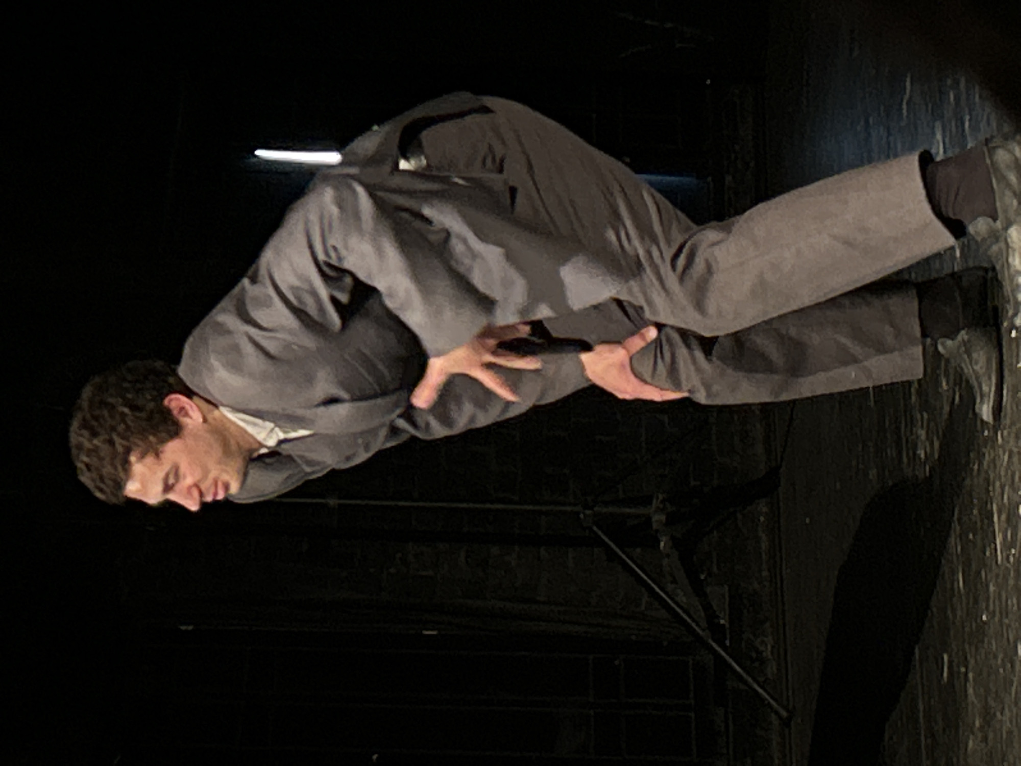 A man wearing a grey suit, white shirt, and black shoes is on a stage in a darkened theater. He is bent over with his hands positioned in front of him. The stage floor appears to be dark and possibly reflective in some areas. There are visible stage props or structures in the background and on the floor near his feet. The location is unknown.