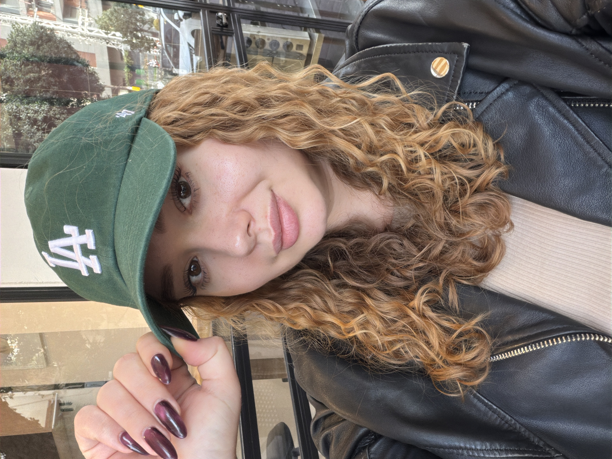 A woman with curly, light brown hair and fair skin is posing for a photo. She is wearing a green baseball cap with a white "LA" logo and a black leather jacket. Her nails are long and painted a dark plum color. The background appears to be an urban environment, possibly a café or restaurant. The location may be Ramat Gan, Israel, based on the prompt, but there are no specific indicators of that location in the image.