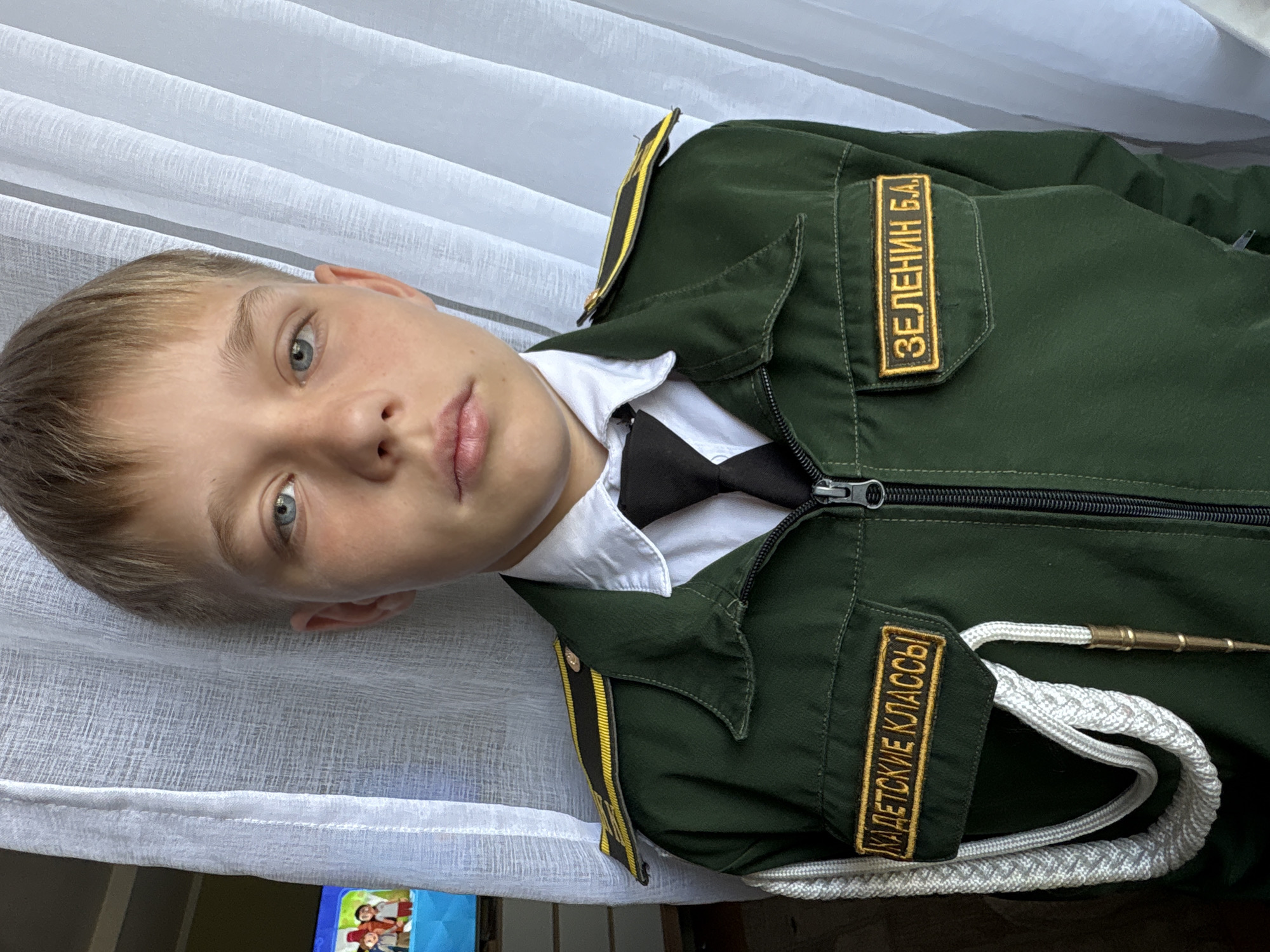 A boy is wearing a green uniform with gold trim and patches, a white shirt, and a black bow tie. The uniform has patches with Cyrillic text. The patch on the right breast reads "ЗЕЛЕНИН Б.А." and the patch on the left breast reads "КАДЕТСКИЕ КЛАССЫ." A white braided rope with a metal tip is draped across the uniform. The boy has fair skin, short blonde hair, and blue eyes. He is looking directly at the camera with a neutral expression. A white curtain hangs behind the boy. The scene appears to be indoors. No other people or specific location within Chaikovsky, Russia can be determined from the image.