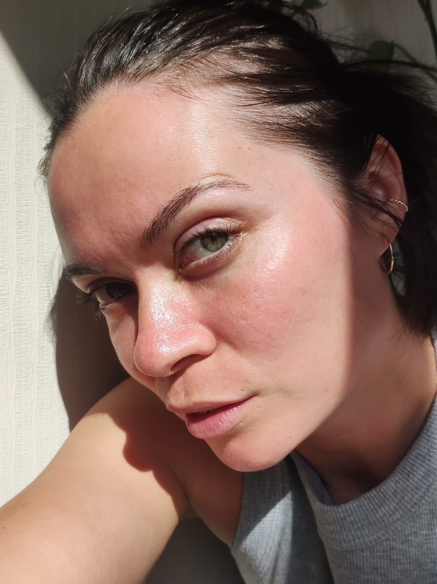 A close-up, sunlit indoor portrait features a person, likely a woman, with fair skin, dark brown hair, and striking green eyes. She is positioned with the left side of her face directly illuminated by strong, natural light, possibly sunlight, which creates prominent highlights on her forehead, nose, and cheeks, and casts a shadow over the right side of her face. The light emphasizes the natural texture of her skin, revealing visible pores, a natural sheen, and some subtle redness, particularly around her nose and cheeks.
Her dark brown hair appears slightly damp or naturally textured, with some strands framing her forehead and temple. Her green eyes are open and directed towards the camera, beneath naturally defined eyebrows. She has delicate eyelashes and there appears to be minimal or no makeup, save for possibly a very light, natural-toned lip color and subtle eye makeup. Her expression is calm and direct, conveying a thoughtful or serene mood.
She is wearing a simple, light gray ribbed top. In her visible right ear, she wears a small hoop earring and another small, twisted earring higher on the ear cartilage. The background to her left is a textured, light-colored wall, while to her right, a darker, out-of-focus element (possibly foliage or a darker interior) is visible. The strong, directional lighting suggests it is daytime. No text is visible in the image. The provided location context is Chaikovsky, Russia.