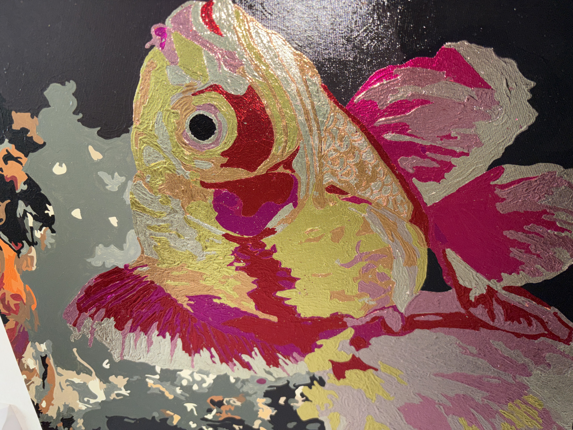 The image is a close-up of a partially completed paint-by-numbers artwork. The subject is a goldfish, predominantly painted in shades of yellow, pink, red, and silver. The eye of the fish is a solid black circle surrounded by red. The background is a uniform black, except for the left side of the image, where portions of an abstract background in shades of gray, orange, and black are present, suggesting other elements of the overall scene are still unpainted. The artwork appears to be on canvas. The location and city (Unknown, Russia) are not discernible from the visual information provided.