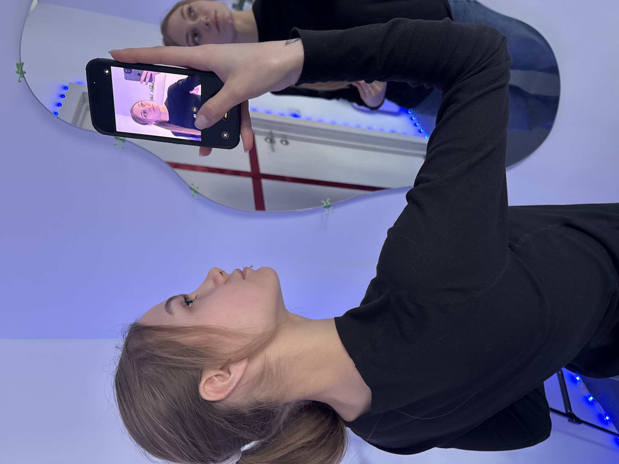 The image displays a young woman taking a mirror selfie, resulting in a recursive "droste effect." In the foreground, the woman is seen in profile, with her head pointing towards the top of the frame, looking upwards. She has light brown hair, which is tied back, and wears a black long-sleeved top. A septum piercing is visible on her nose.
Her reflection is prominently featured in a large, irregularly shaped mirror that dominates the background. In the reflection, she holds a black smartphone with her right hand, aiming it towards the mirror to capture the image. The phone's screen itself shows her reflection, further depicting her holding the phone, thus creating an infinite series of reflections within reflections. Visible on the phone's screen are mode indicators like "PHOTO" and zoom options such as "0.5x," "1x," and "2x."
The room is bathed in a distinct blue and purple light, originating from an LED strip that runs along a surface behind the mirror and is also reflected in it. Several small, bright green, frog-shaped decorative items are scattered across the mirror's surface. The scene takes place indoors, characterized by its modern and artificially lit ambiance.