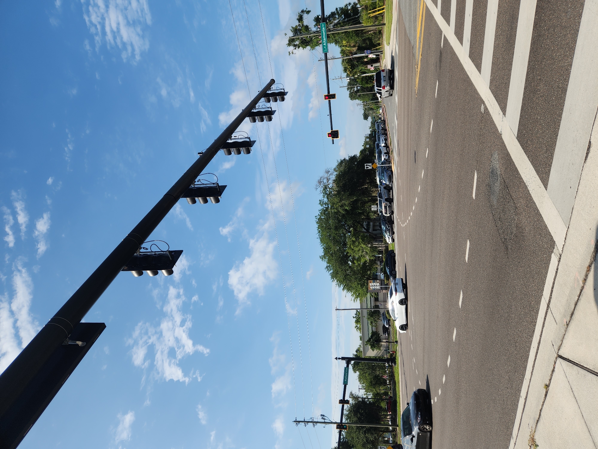 This is a rotated, medium-angle shot of a street in Altamonte Springs, Florida. The scene depicts a road with multiple lanes and a pedestrian crossing marked by white stripes. Several cars are parked along the right side of the road, and others are in traffic. The sky is a bright blue with scattered white clouds, suggesting it's daytime and the weather is clear.
Dominating the left side of the image is a tall, black traffic signal pole with multiple traffic lights. In the distance, another traffic signal pole is visible, displaying red lights. A street sign with green lettering reads "E Altamonte Dr". Trees and buildings line the right side of the road. The overall impression is of a typical suburban street scene on a sunny day. There are no people visible in the image.