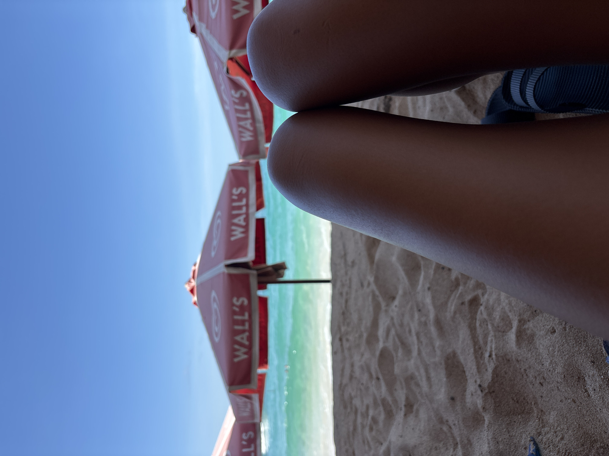 A vertically oriented shot depicts a beach scene in Kuta Selatan, Indonesia. The foreground is dominated by the lower limbs of one or more individuals on the sand, with part of a blue shoe visible. Behind these, a row of identical red beach umbrellas with the "WALL'S" logo stretches back. Beyond the umbrellas, the turquoise ocean meets the horizon under a clear blue sky. The scene suggests a sunny day at the beach with people enjoying the view and weather.