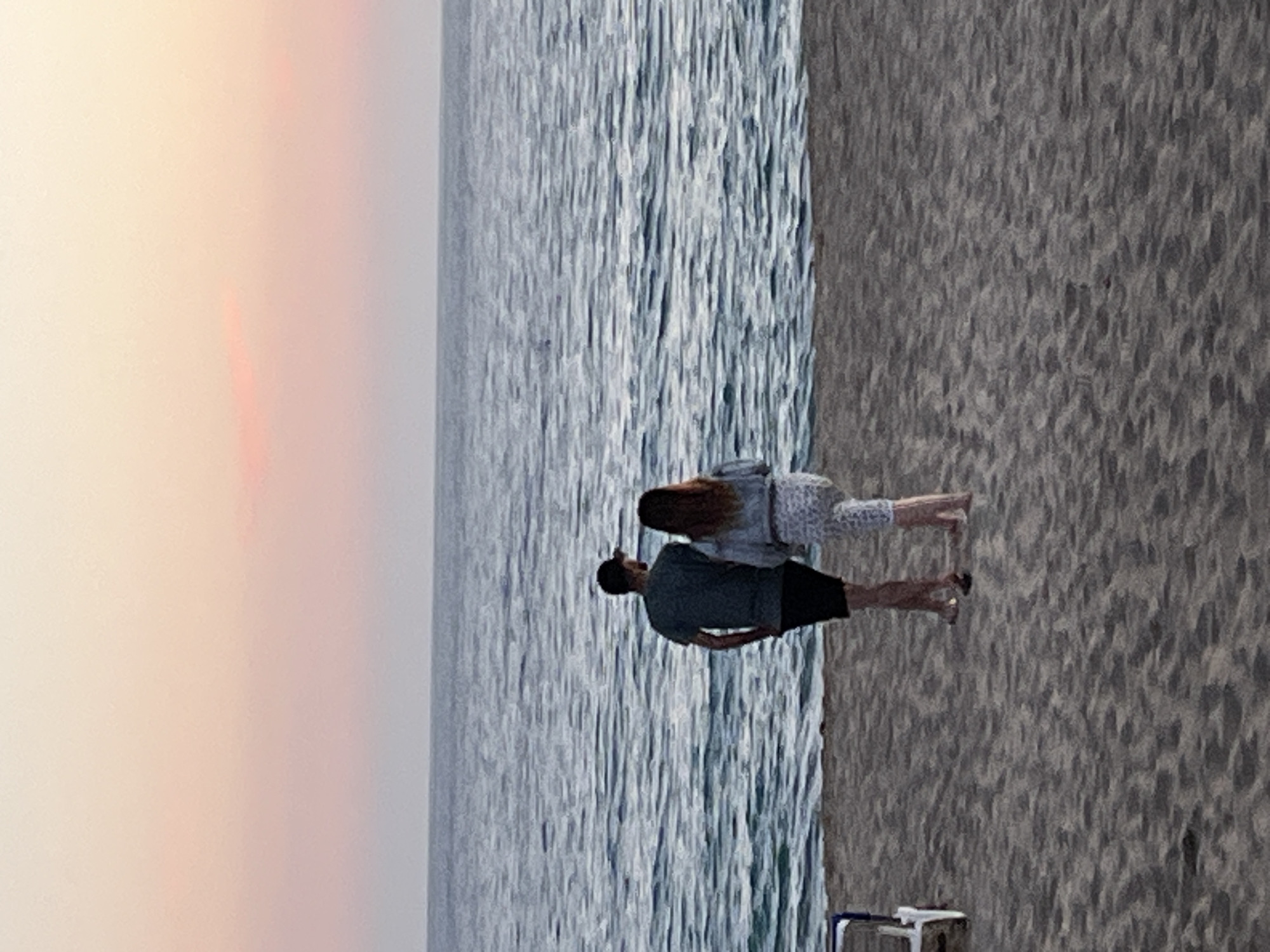 A couple stands on a sandy beach next to the sea. The woman has long brown hair and is wearing a patterned outfit. The man has short dark hair and is wearing a dark T-shirt and dark shorts. The sky is light-colored, suggesting either sunrise or sunset. The water is rippled, and there is some sort of structure near the lower right corner. The location is Alanya, Turkey.
