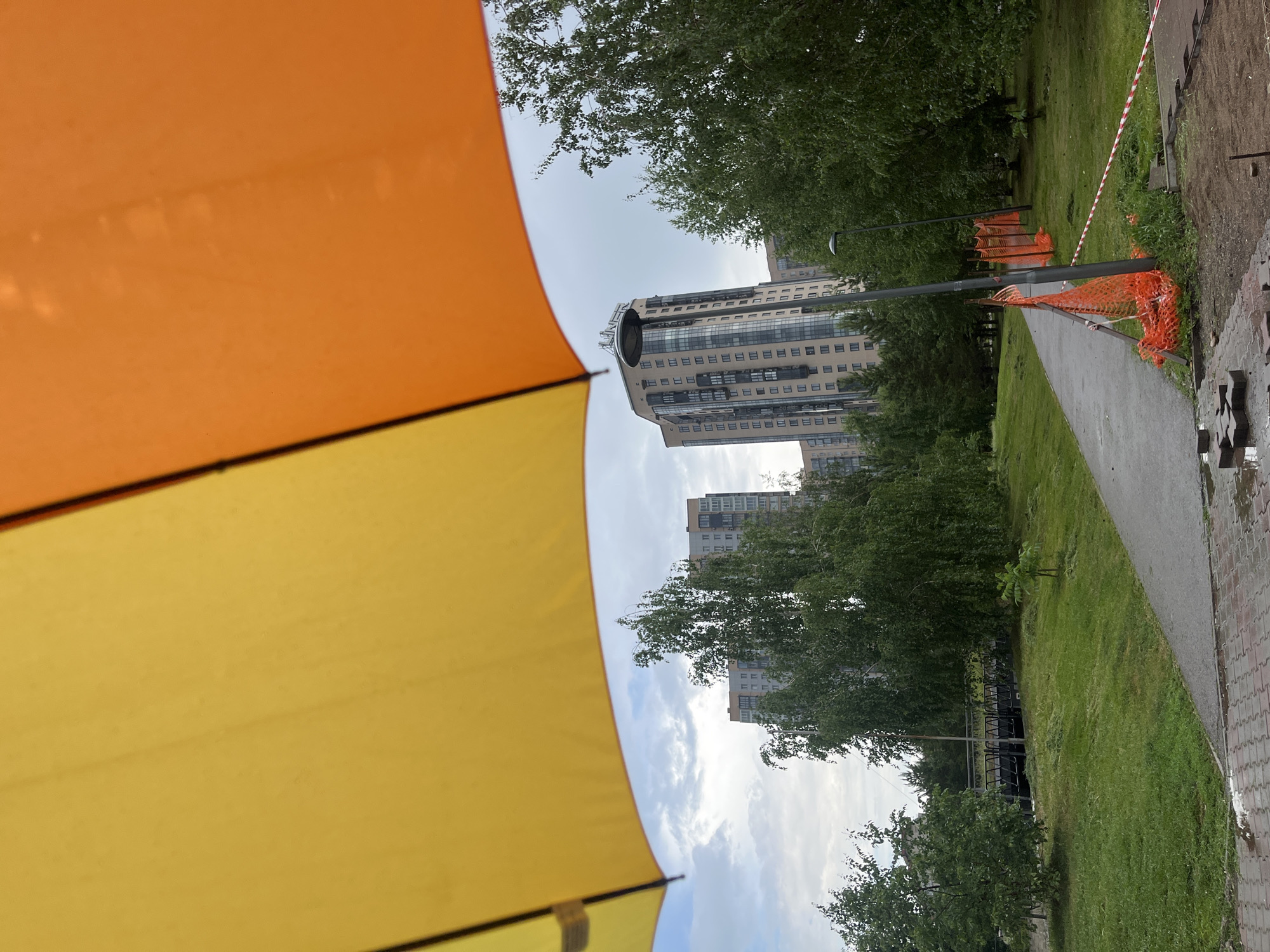 The image depicts an outdoor scene with a bright yellow and orange umbrella dominating the foreground, partially obscuring the view. Beyond the umbrella, a pathway cuts through a grassy area with trees on either side. In the background, several modern apartment buildings stand tall against a cloudy sky. Construction fencing, made of orange netting and white and red tape, is visible along the pathway, indicating some ongoing work. A bridge can be seen in the middle ground, crossing a body of water. The ground near the pathway is paved with bricks, some of which are wet and appear to have been recently disturbed. The scene suggests a residential area in Krasnoyarsk, Russia, possibly a park or recreational space undergoing renovation.