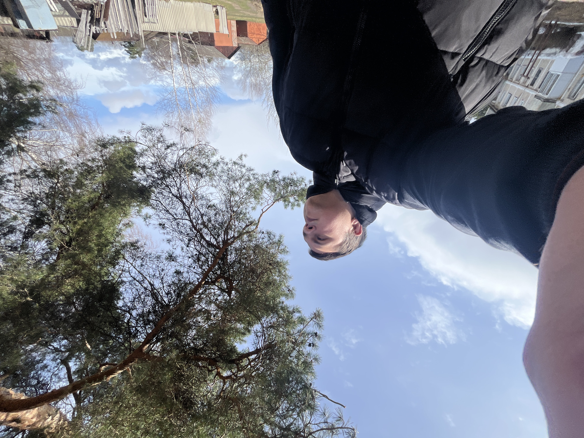 The image is an upside-down, low-angle selfie of a person wearing a dark jacket. The person's head and upper body are in the center, with a view of the sky and trees in the background. The background also shows a blurry landscape of buildings, some with corrugated metal roofs, suggesting an area in the vicinity of Razdolye, Russia.