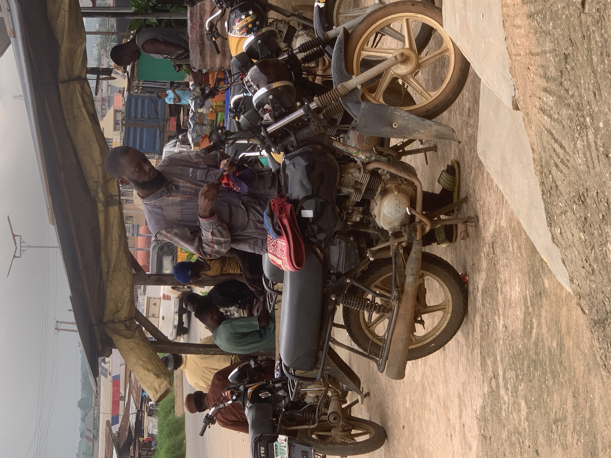 The image depicts an outdoor scene in Shagamu, Nigeria, likely a marketplace or roadside gathering area. In the foreground, two motorcycles are prominently featured. The one on the left is black with a license plate reading "OGUN 591-VJ". The motorcycle on the right is partially obscured, with a yellow section and stars visible on its front.
A man, dressed in a grey plaid shirt and dark pants, is standing and engaged with a red and blue cloth in his hands, possibly performing a repair or maintenance task on one of the motorcycles. He has a beard. To his right, another man, wearing a grey sleeveless shirt, stands looking down.
In the background, several other individuals are present. One person in a green shirt is seated. Further back, under a makeshift canopy supported by poles, other people are visible, including one wearing a blue hat and another in a yellow vest. A white pickup truck and a blue truck are also visible in the background, suggesting a busy commercial area. The environment appears dusty, with a rough, sandy ground. Buildings with corrugated metal roofs and some vegetation are present in the distance, indicating an urban or semi-urban setting. The sky is overcast, contributing to a muted color palette.