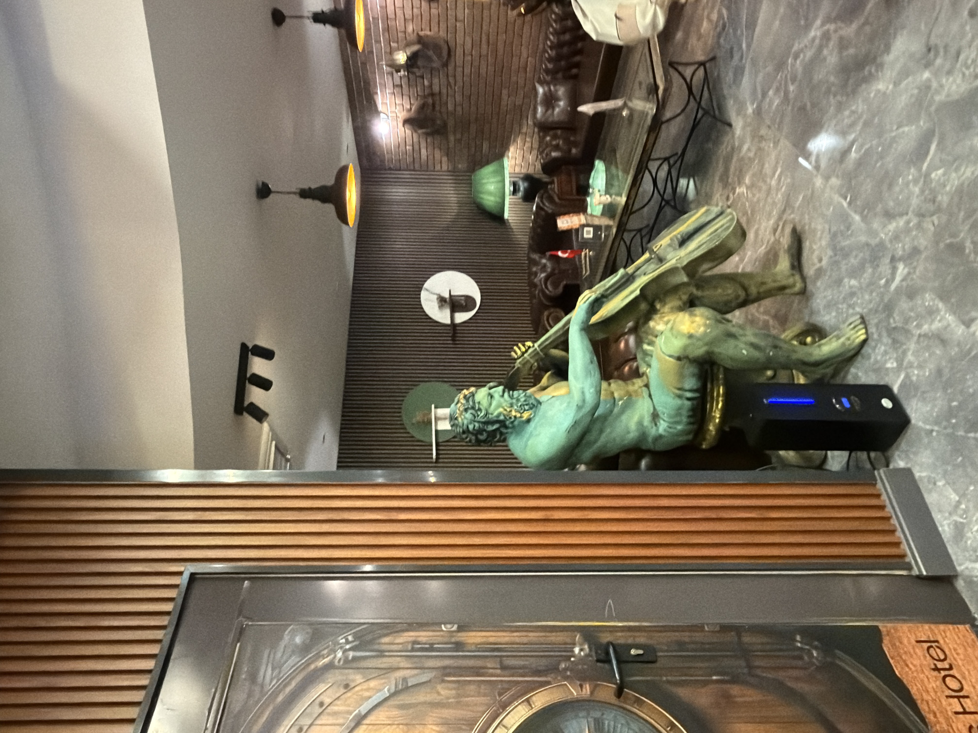 The image depicts an elaborately decorated indoor space, likely a hotel lobby or lounge, showcasing an eclectic design style. The dominant subject is a large, verdigris-patinated statue of a muscular, bearded male figure, possibly a sea god or mythical character, reclining or semi-sitting on a low platform. The figure holds a spear or trident-like object. Beside the statue, a black rectangular device with a blue illuminated strip is visible.
The walls feature various textures: a light-colored upper section, a middle section with horizontal wooden slat paneling, and a brick wall further in the background. Mounted on the brick wall are several dark, indistinct animal-like figures, possibly taxidermy or statues. The ceiling is light-colored, from which multiple pendant lights with a warm, orange glow hang, creating an atmospheric, dimly lit environment. Additional wall-mounted spotlights are also present. The floor is composed of polished, mottled gray and brown stone or tile, reflecting light sources and architectural elements.
In the lower foreground, a decorative door or panel with wooden and metallic details, including a porthole-like design, is partially visible. Text on this element indicates the location, reading "...Hotel." No human activity is depicted in the static scene.