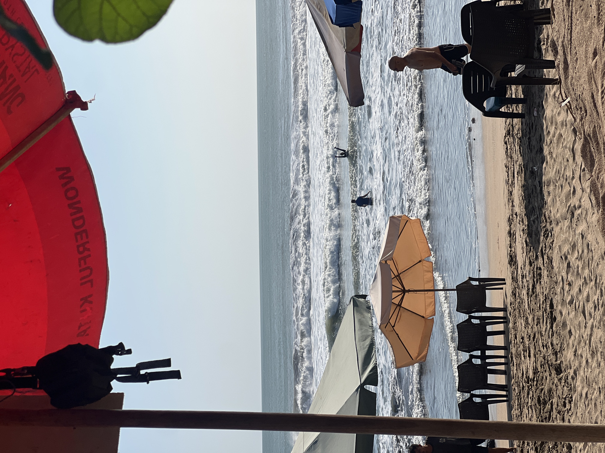 A wide shot of Kuta Beach in Indonesia shows a sunny day with people enjoying the water and the sand. In the foreground, several empty black plastic chairs are arranged on the sandy beach, some under a light orange beach umbrella. To the left, a dark wooden pole supports a red canopy with white lettering that appears to be upside down. In the middle ground, the ocean has moderate waves, and several people are in the water. One person is standing with their arms raised, while another, wearing a blue shirt, walks in the surf. In the background, the ocean stretches to the horizon under a clear blue sky. A man in dark swim trunks is walking along the water's edge in the right middle ground. To the left of the frame, a person in dark clothing is partially visible. Two large, dark green beach umbrellas are visible further back on the sand.