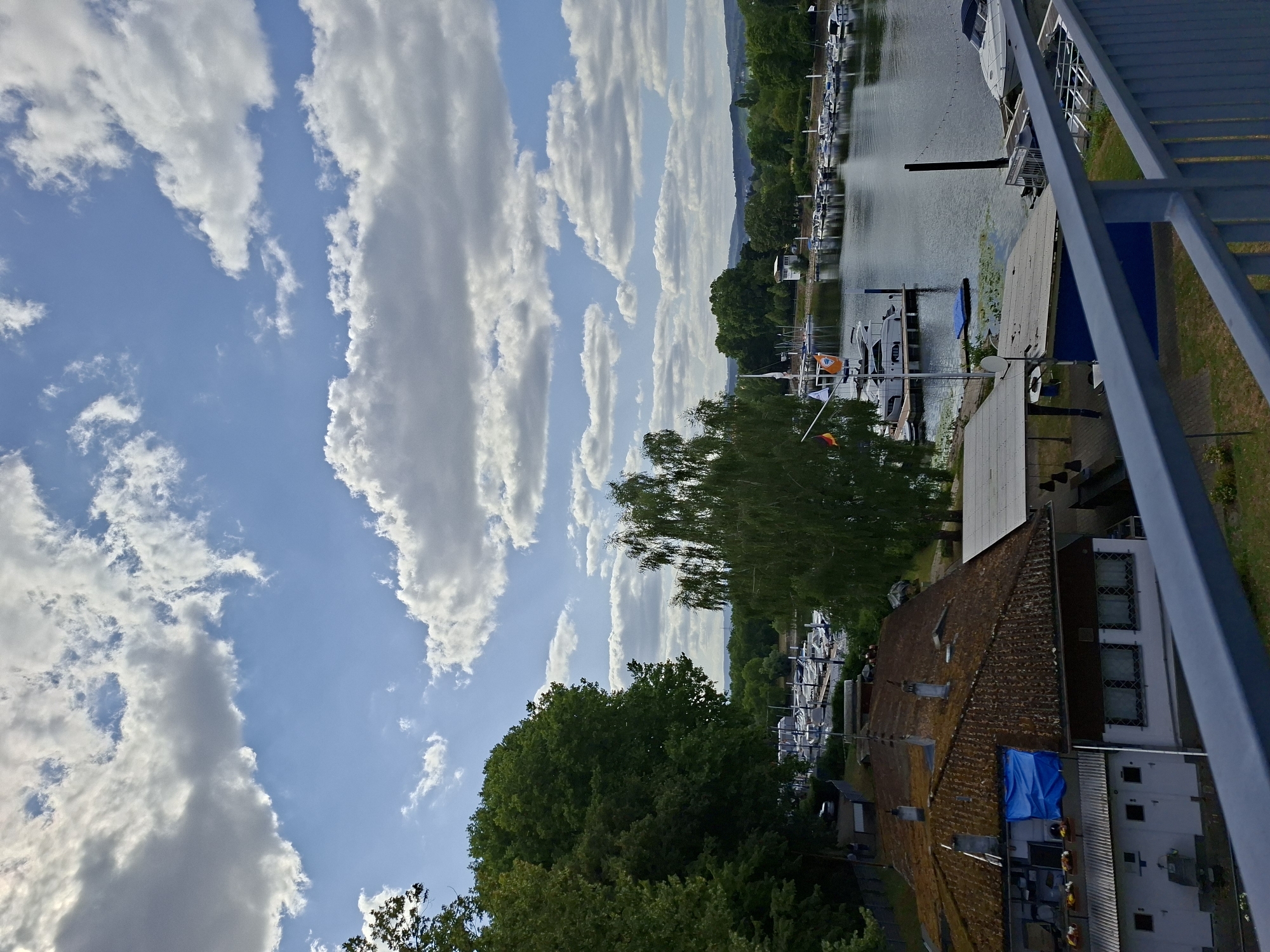 The image depicts a view of the Rhine river in Wiesbaden, Germany, on a cloudy day. In the foreground, a railing of a bridge or walkway is visible, partially obscuring a view of buildings and trees. The buildings appear to be residential or commercial, with tiled roofs and some with blue tarp coverings. Further in the background, the Rhine river is lined with numerous boats docked at a marina. The water is calm, reflecting the sky. Trees and greenery flank the riverbanks, and in the distance, rolling hills can be seen. The sky is characterized by a mix of blue patches and large, fluffy white clouds. There are no discernible people or animals in the scene.