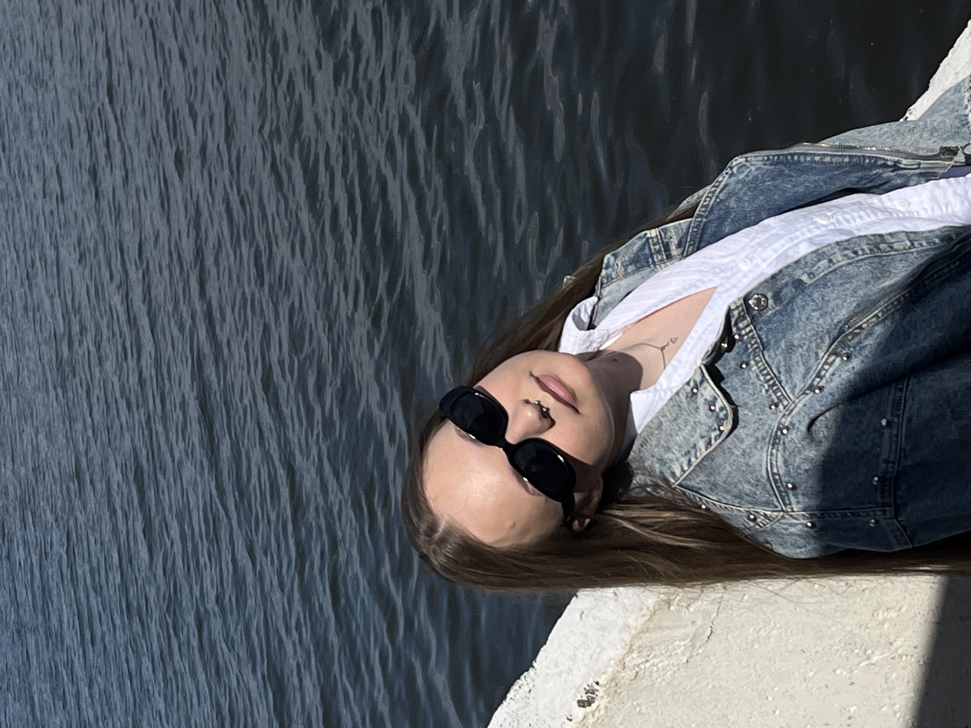 A woman is lying on a concrete ledge next to a body of water. The woman is wearing sunglasses, a white shirt, and a denim jacket. The body of water is possibly a lake. The location is likely Chaikovsky, Russia.