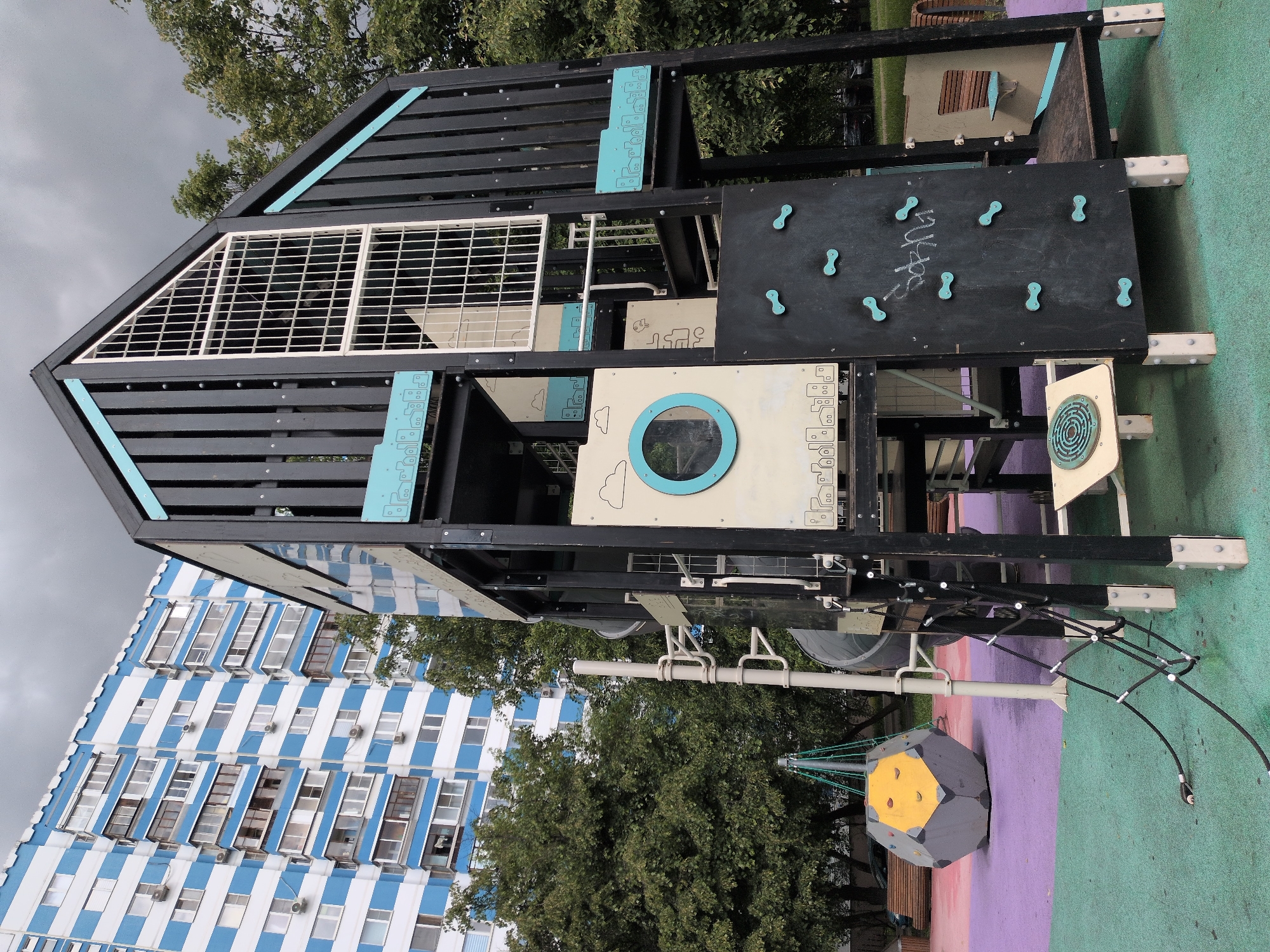 The image shows a children's playground structure in a city, likely Moscow, Russia. The playground equipment features a climbing wall, a rope bridge, and various decorative panels with geometric shapes and designs. The structure is predominantly black with light blue accents.
In the background, there is a tall apartment building with a distinctive blue and white striped pattern. The sky is overcast. Trees surround the playground, adding to the urban setting. The ground is covered in a multi-colored surface.