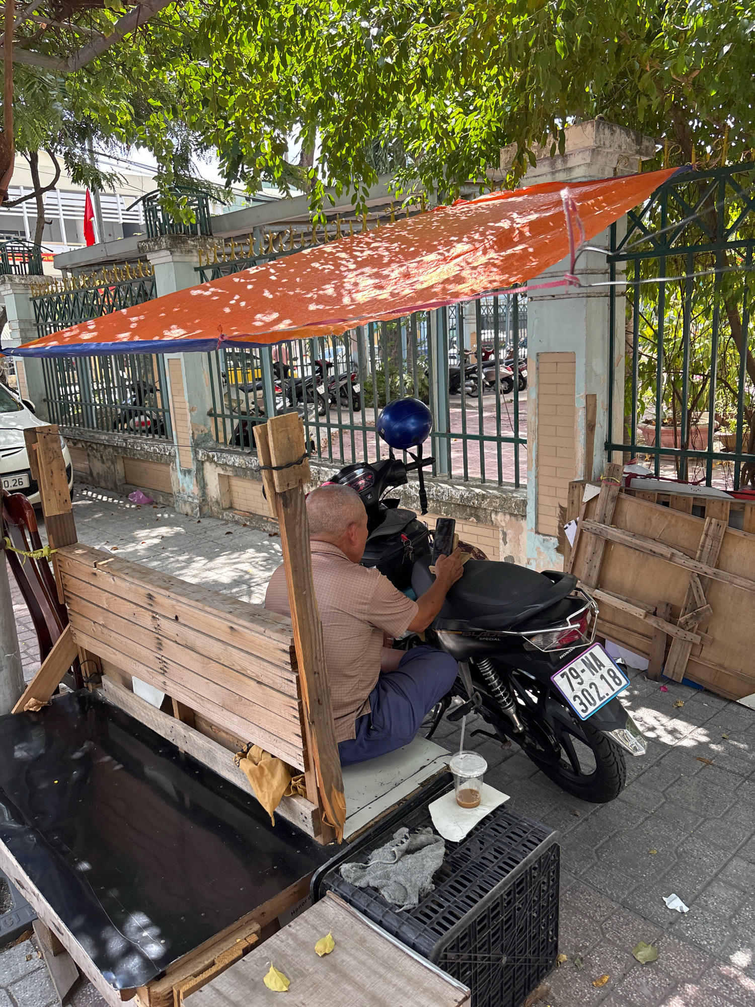In Nha Trang, Vietnam, an elderly man is seated on a motorbike under a makeshift orange tarp shelter. He is looking at his phone, holding it in his left hand. The motorbike, a black Honda, is parked next to a rustic wooden bench. In front of the bench, a dark, reflective surface, possibly a table, is visible. A black plastic crate is on the ground to the right of the bench, holding some items. A clear plastic cup with a beverage sits on a white paper on the crate. The scene appears to be a street-side setup, with a metal fence in the background and trees providing shade. The pavement is paved with grey tiles.