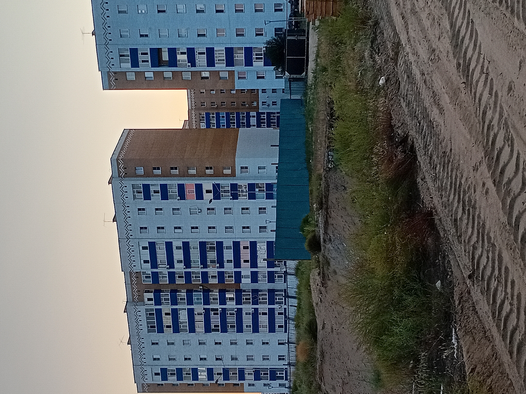 The image shows two tall residential buildings with light blue and white facades, accented with dark blue horizontal bands. The buildings have multiple stories, each with numerous rectangular windows. Decorative triangular patterns are visible along the rooftops of both buildings. In the foreground, there is a dirt embankment with sparse green vegetation. A textured dirt path with tire tracks runs through the bottom of the frame. A turquoise metal fence is situated in front of the buildings. The sky is clear and pale, suggesting either dawn or dusk. The scene appears to be a typical residential area in the city of Semey, Kazakhstan, likely depicting a new or under-construction development given the unfinished appearance of the foreground. There are no people or animals visible in the image.
