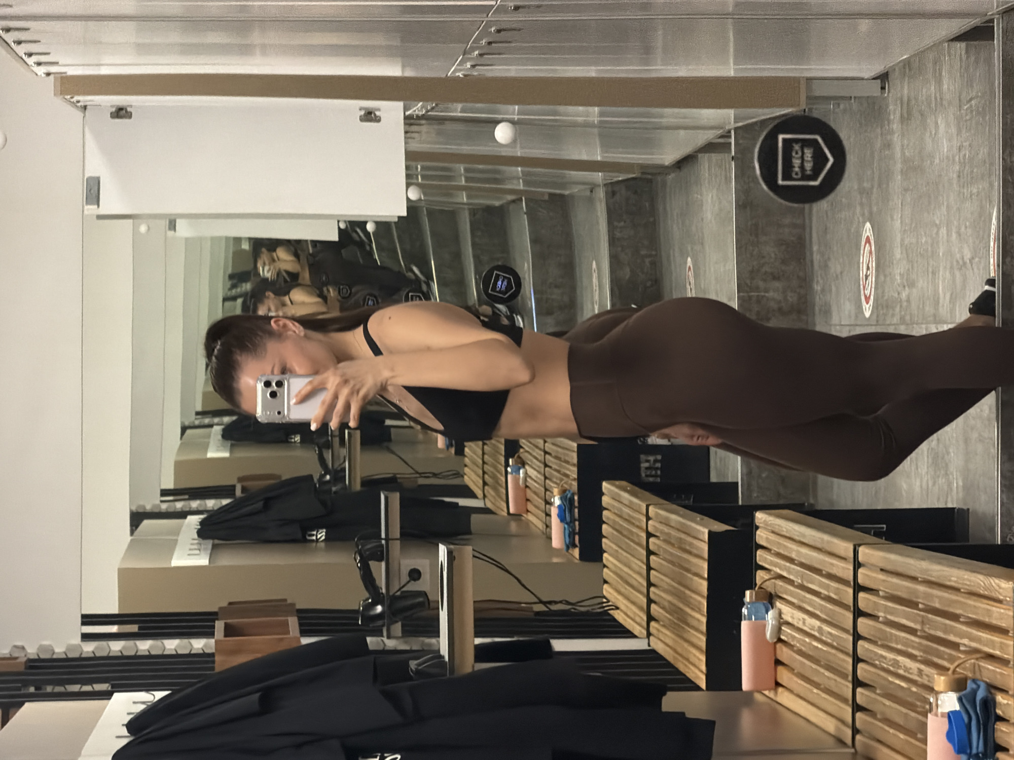 A woman is taking a mirror selfie in what appears to be a fitting room or changing area of a retail store. She is wearing a black sports bra and brown leggings. The background shows a gray and white interior with fitting room stalls, wooden dividers, clothing racks, and a "Check Here" sign. The city and country are not evident from the image.