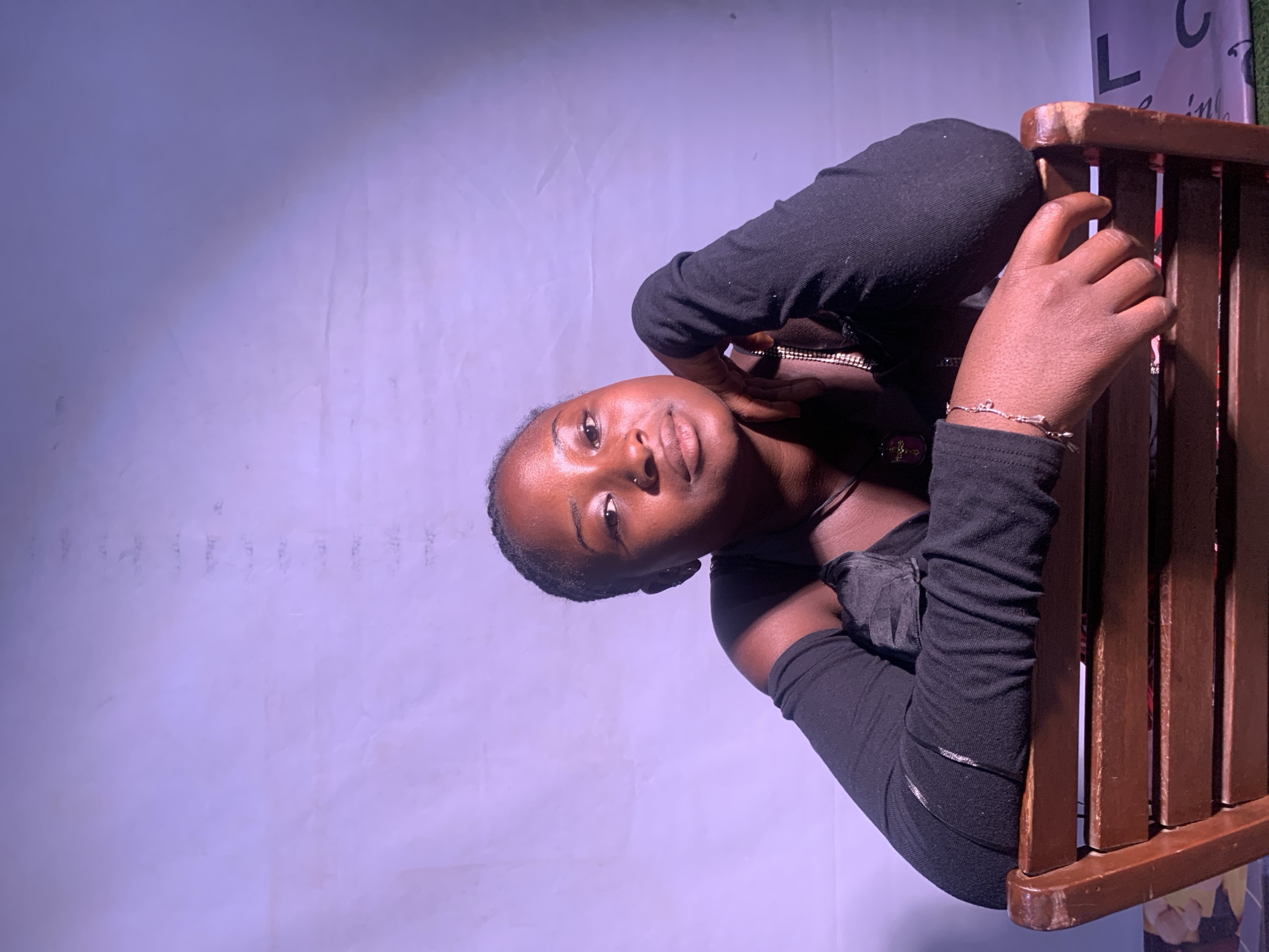 A young woman is seated in a wooden chair, with a backdrop of what appears to be a white wall. She is wearing a long-sleeved black top and a silver bracelet on her right wrist. Her right hand is resting on the chair. The setting appears to be indoors, likely in the city of Jos, Nigeria. More detail is required to ascertain specifics about the location within the city of Jos, Nigeria.