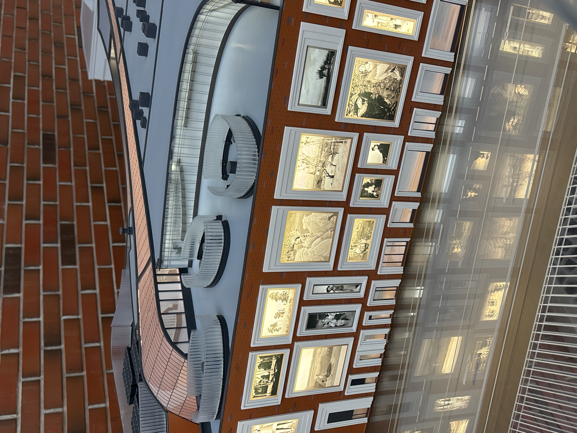 The image shows a section of the Tretyakov Gallery building in Moscow, Russia, featuring a facade with numerous framed photographs and architectural embellishments. The facade is adorned with a series of light fixtures shaped like segmented arcs and a pattern of square framed images that each contain some image inside. The reflections on the windows create repeating imagery, including the sky. The background shows the texture of a brick wall.