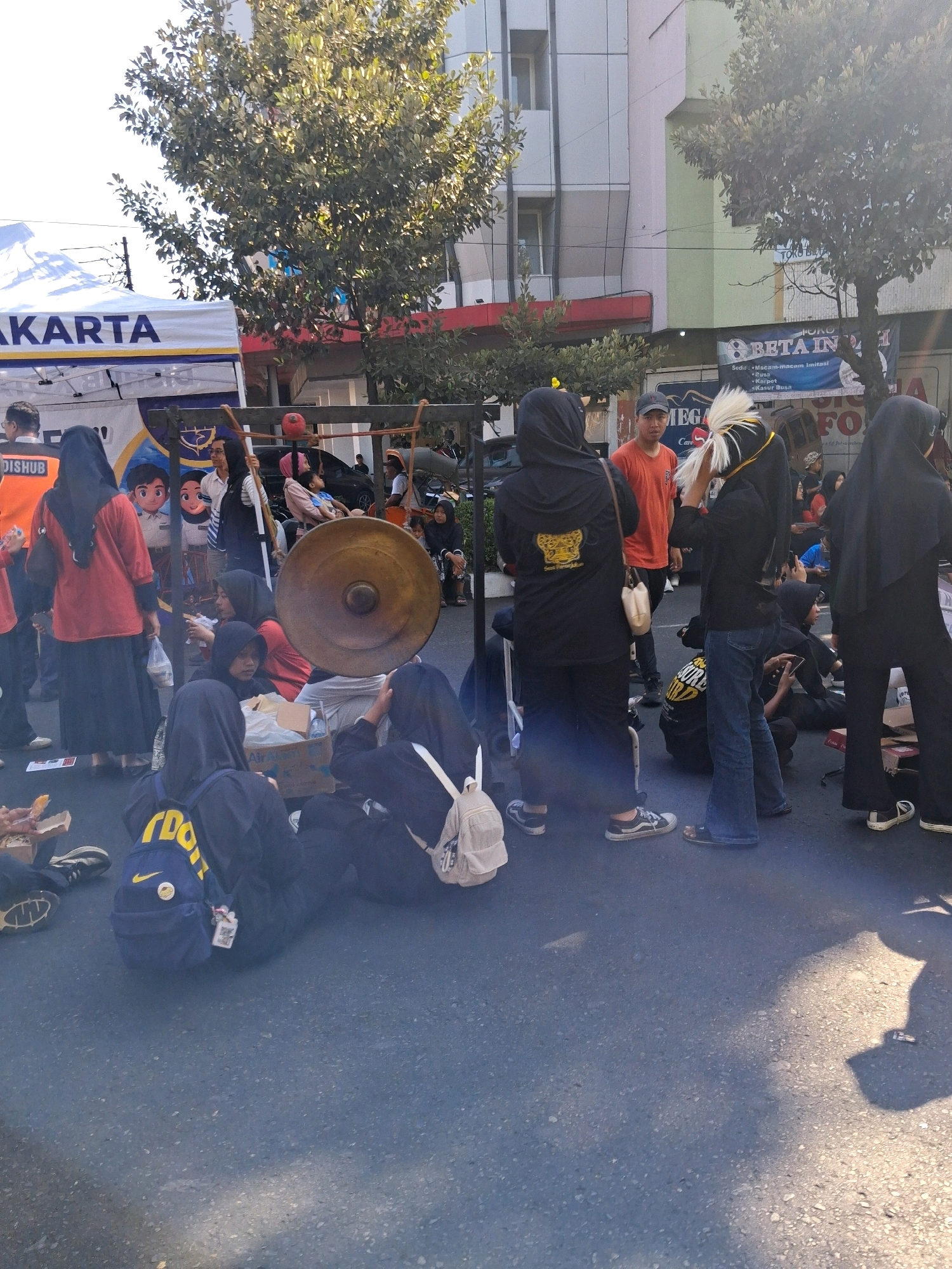 The image depicts an outdoor scene, likely a street or public square, in Surakarta, Indonesia. A group of people, primarily young women wearing hijabs and long clothing, are gathered. Some are sitting on the ground, while others stand. In the foreground, a young woman wearing a black hijab and a yellow backpack with the word "DOJO" printed on it sits on the ground, facing away from the camera. To her right, another young woman in a black hijab is also seated, her back to the camera, with a white backpack beside her. A large, circular brass gong, suspended from a wooden frame, is a prominent object in the middle ground, suggesting a cultural or performance event. Several individuals are positioned around this instrument. In the background, buildings with a mix of modern and traditional architecture are visible, along with trees. Signs in Indonesian are present, including one that reads "JAKARTA". The overall impression is of a communal gathering, possibly related to a local festival or cultural activity.