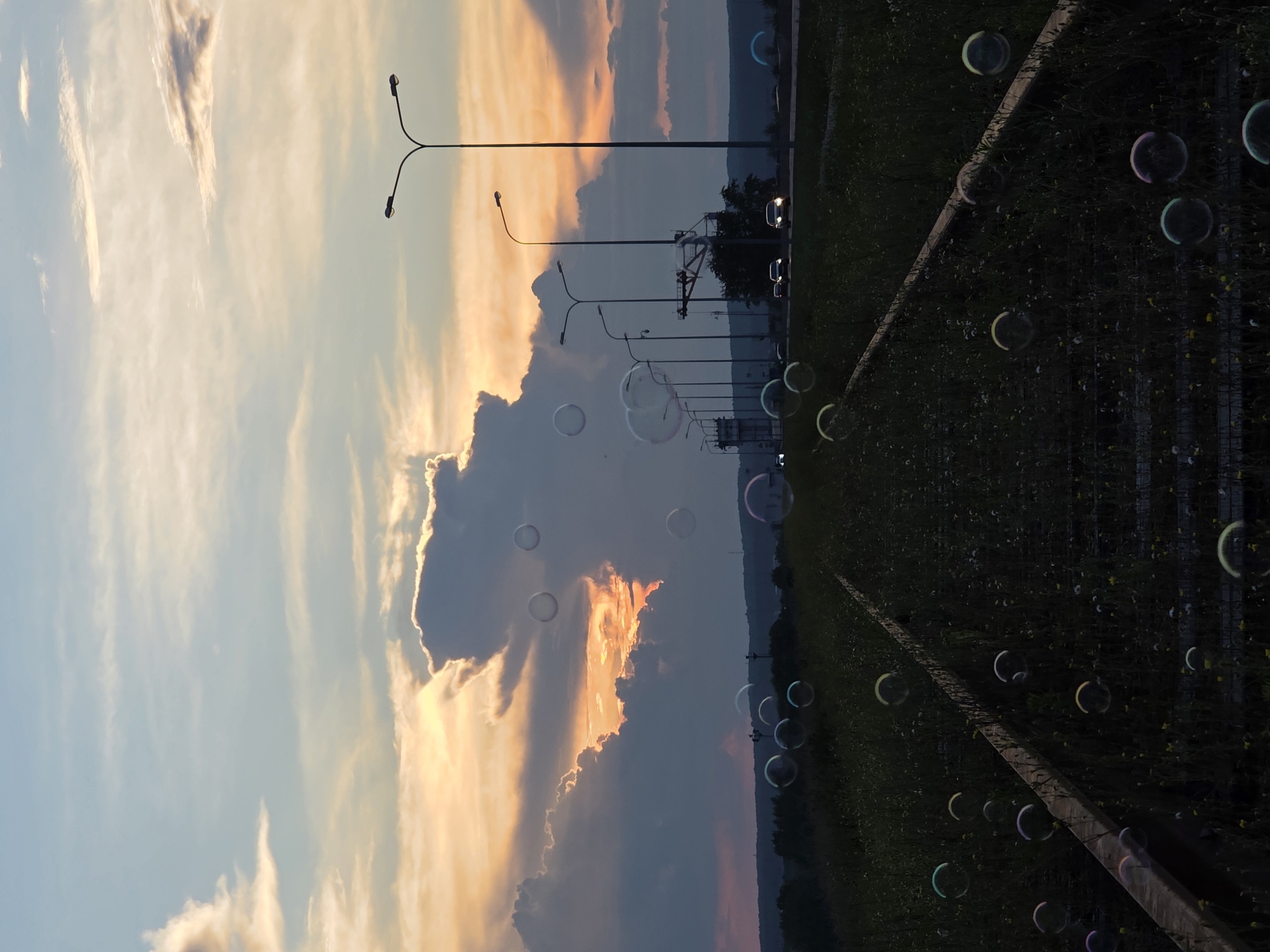 An eye-level shot captures a dusky sunset over a grassy embankment with railroad tracks leading towards a highway. The sky is painted with vibrant orange and yellow hues, contrasting with dramatic dark clouds. Numerous soap bubbles drift across the frame, adding a whimsical touch to the scene. The highway, lined with streetlights, features a few cars in motion, their headlights illuminating the road. In the distance, a forested hillside forms the horizon. The image appears to have been taken near the Chaikovsky City Hospital in Chaikovsky, Russia, given the specific mention of this location in the provided context, though no specific landmarks of the hospital are visible.
