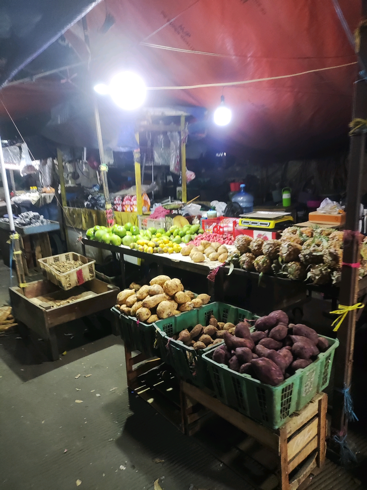 An outdoor market stall is illuminated by bright overhead lights. The stall displays a variety of produce. In the foreground, there are two green plastic crates filled with dark purple sweet potatoes and lighter brown sweet potatoes. Behind these, there is a wooden crate filled with what appears to be ginger. Further back, a table is laden with green apples, yellow citrus fruits, and red radishes. To the right of the produce table, a stack of pineapples is visible. In the background, there are shelves with packaged goods, a water cooler, and a yellow scale. The market is covered by a red tarp. The ground is paved and appears to be wet in places, with some scattered debris. The scene suggests a typical market setting in East Jakarta, Indonesia, likely operating at night or in low-light conditions. There are no people visible in the image.