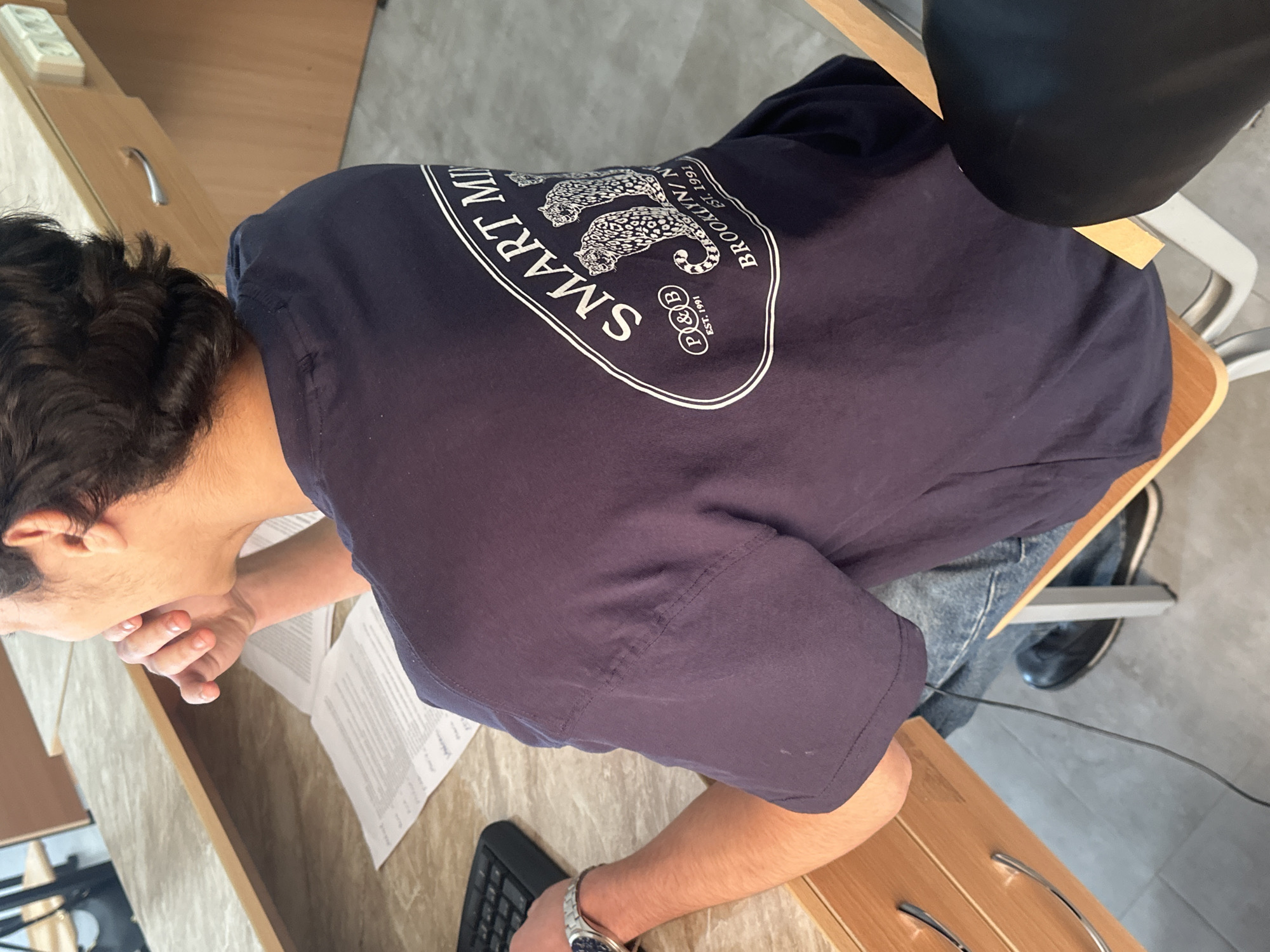 A person wearing a dark blue shirt with a white logo that reads "SMART MIND BROOKLYN NY EST. 1991" is seated at a desk. The logo also features two leopard-like figures. The individual is positioned facing towards a surface covered in papers. A keyboard and a watch-adorned wrist are visible on the desk. A black cylindrical object, potentially a lamp or speaker, is visible in the upper right of the frame, partially obscured. The person is sitting on a chair with a wooden seat. The location in the city is unknown.