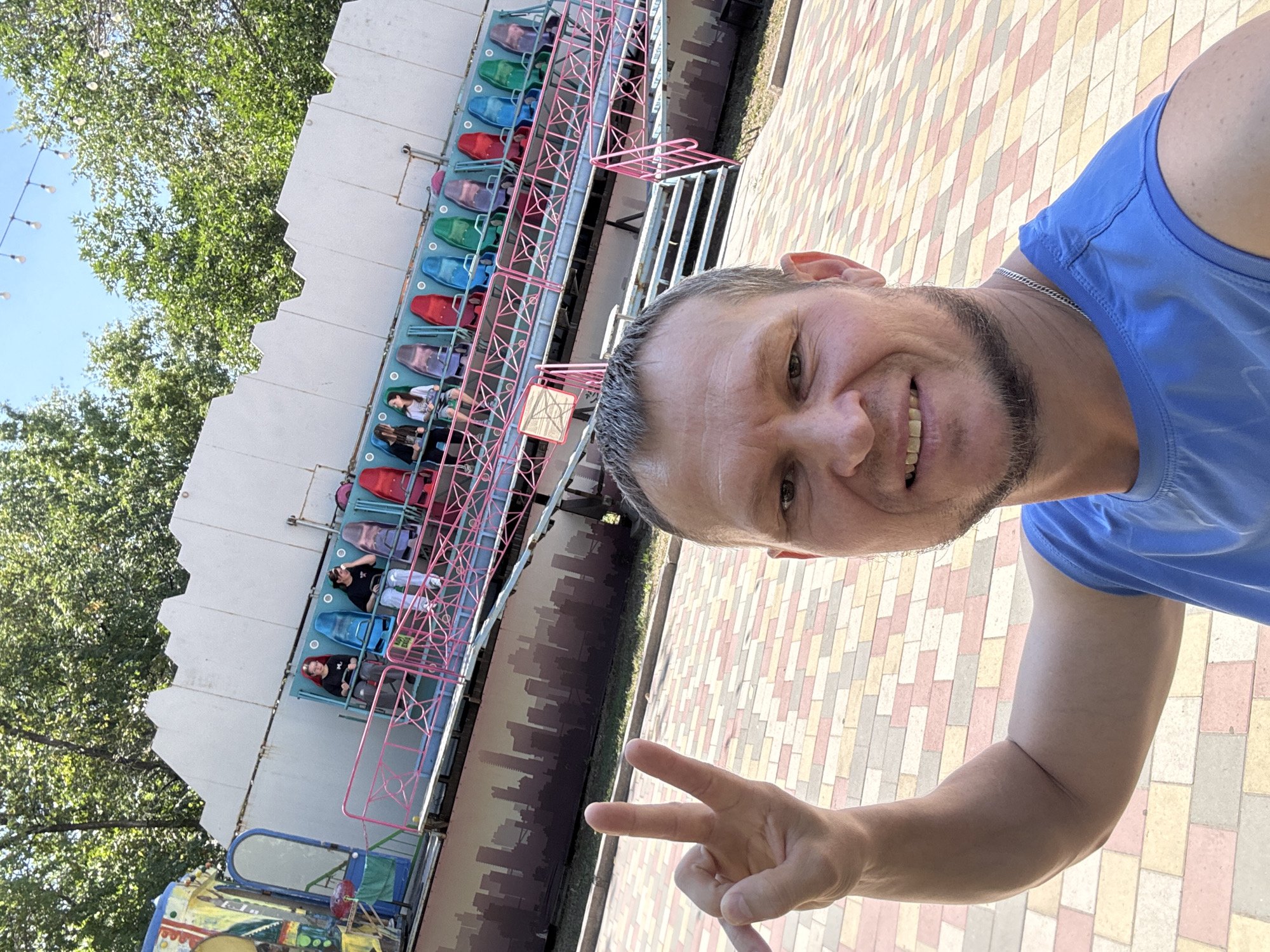 A man in a blue t-shirt takes a selfie in front of an amusement park ride. He is holding up two fingers in a peace sign. The ride is colorful with rows of seats, and several people are visible on it. The ground is paved with interlocking bricks. Trees and a bright blue sky are in the background. The image was taken in Oskemen, Kazakhstan.