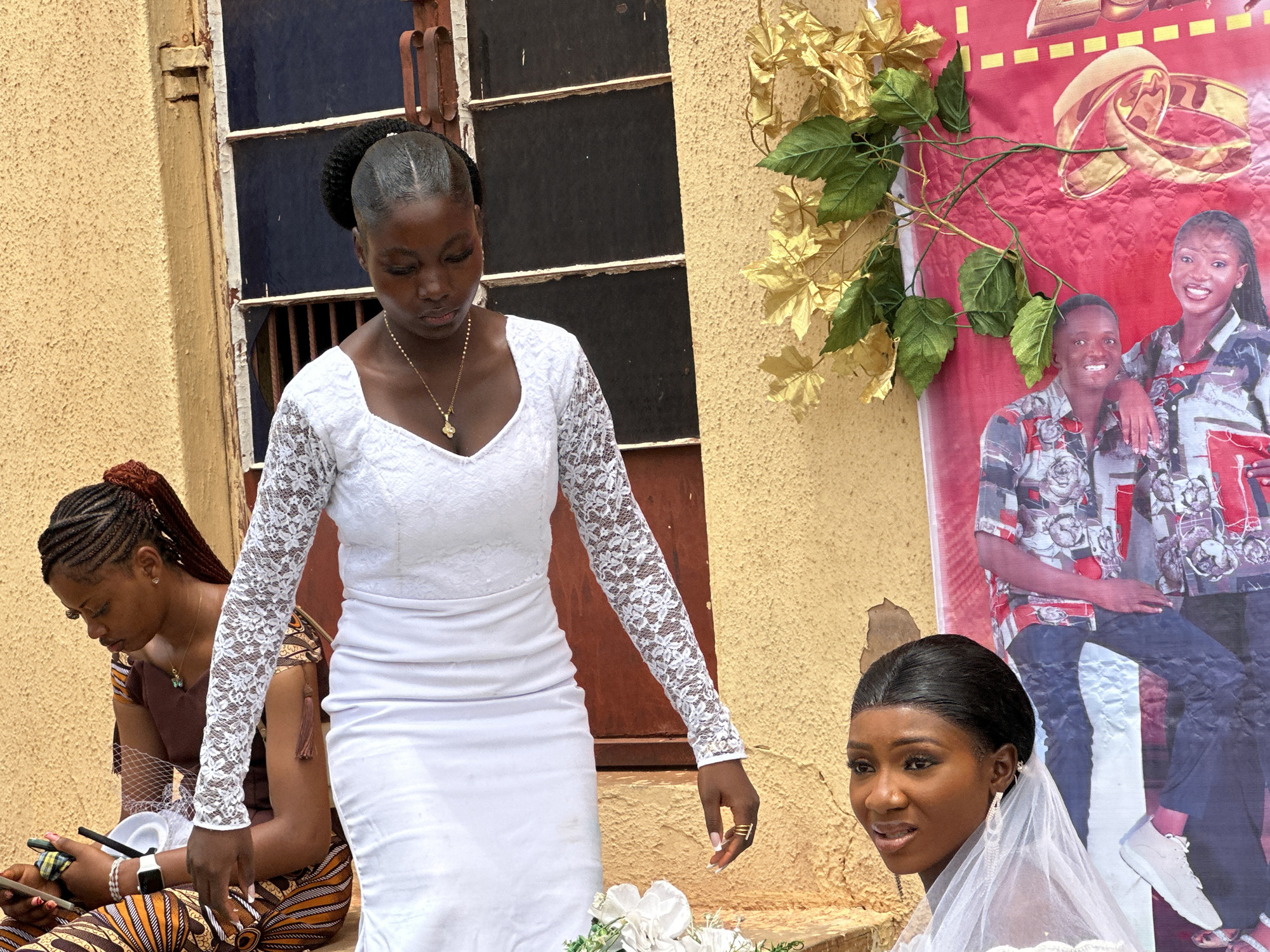 Captured outdoors in an unknown city, the image features a wedding scene against a yellow building. A woman, likely the bride, stands in a white, lace-sleeved dress, looking down with a necklace visible. To her left, a seated woman in patterned clothing uses a mobile phone. To the right, another woman, possibly a bridesmaid, is partially visible in a white dress and veil, gazing to the left. A wedding banner with a photograph of a couple and golden rings decorates the wall behind them, enhanced by faux golden foliage. A weathered window with a brown frame is also visible.
