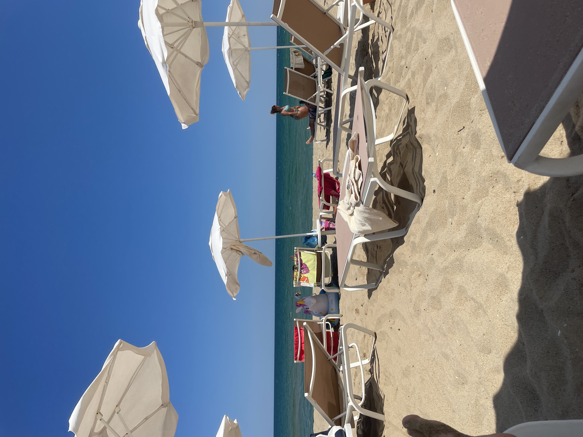 This is a scene of a sunny beach in Obzor, Bulgaria. The foreground is dominated by sand and several white beach chairs with beige cushions. In the middle ground, more beach chairs and umbrellas are visible, along with some beach towels and a large inflatable unicorn. Further back, the calm sea stretches out to the horizon under a clear blue sky. A woman in a turquoise bikini is visible in the distance, interacting with a person lying on a lounge chair. The overall impression is one of a peaceful and leisurely day at the seaside.