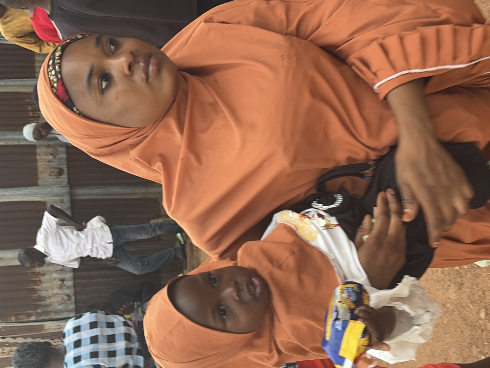 In an outdoor, urban environment, a Black woman wearing an orange hijab and an orange garment holds a young Black child, also in an orange hijab. The child is wearing a white garment with colorful cartoon characters. The woman's hands are around the child, and the child is holding a blue and yellow object. In the background, a Black man in a white collared shirt and blue jeans stands with his hand on his hip, facing away from the camera. Behind him, a corrugated metal fence runs horizontally. Other individuals are partially visible in the background, including a person in a yellow shirt and another in a red shirt. The ground is covered in dirt.