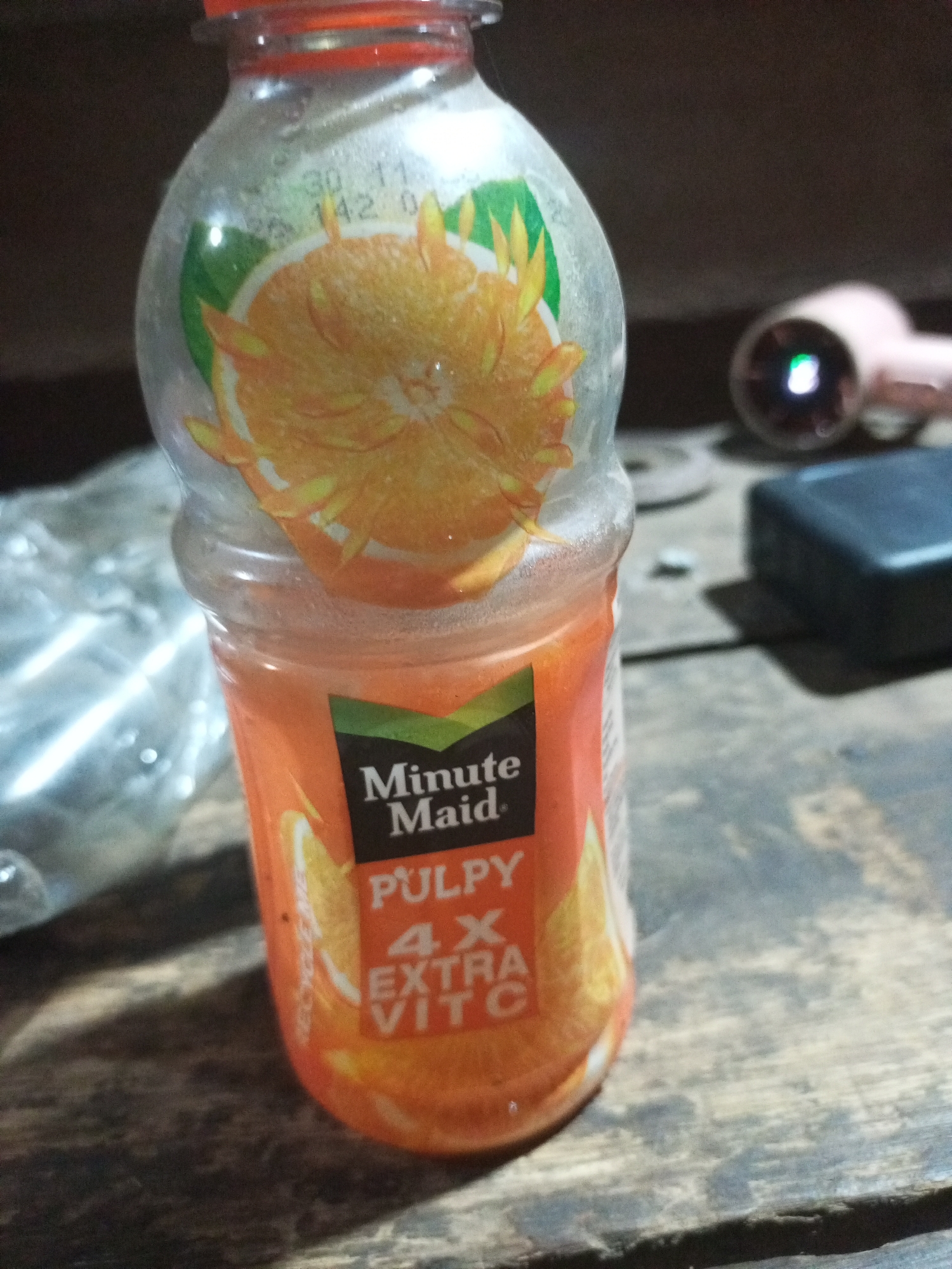 A clear plastic bottle of Minute Maid Pulpy Orange juice with a red cap is shown in the foreground. The bottle is partially filled with orange liquid and has a label that reads "Minute Maid Pulpy 4X Extra Vit C". The bottle is placed on a weathered wooden surface. In the background, to the right of the bottle, a pink object with a green light emanating from it is visible, possibly a flashlight or electronic device. A dark, rectangular object is also present behind the bottle. To the left of the bottle, a blurred image of a shiny, reflective surface, potentially plastic wrapping, is visible. The overall environment appears to be indoors, with a dark background suggesting a dimly lit setting.