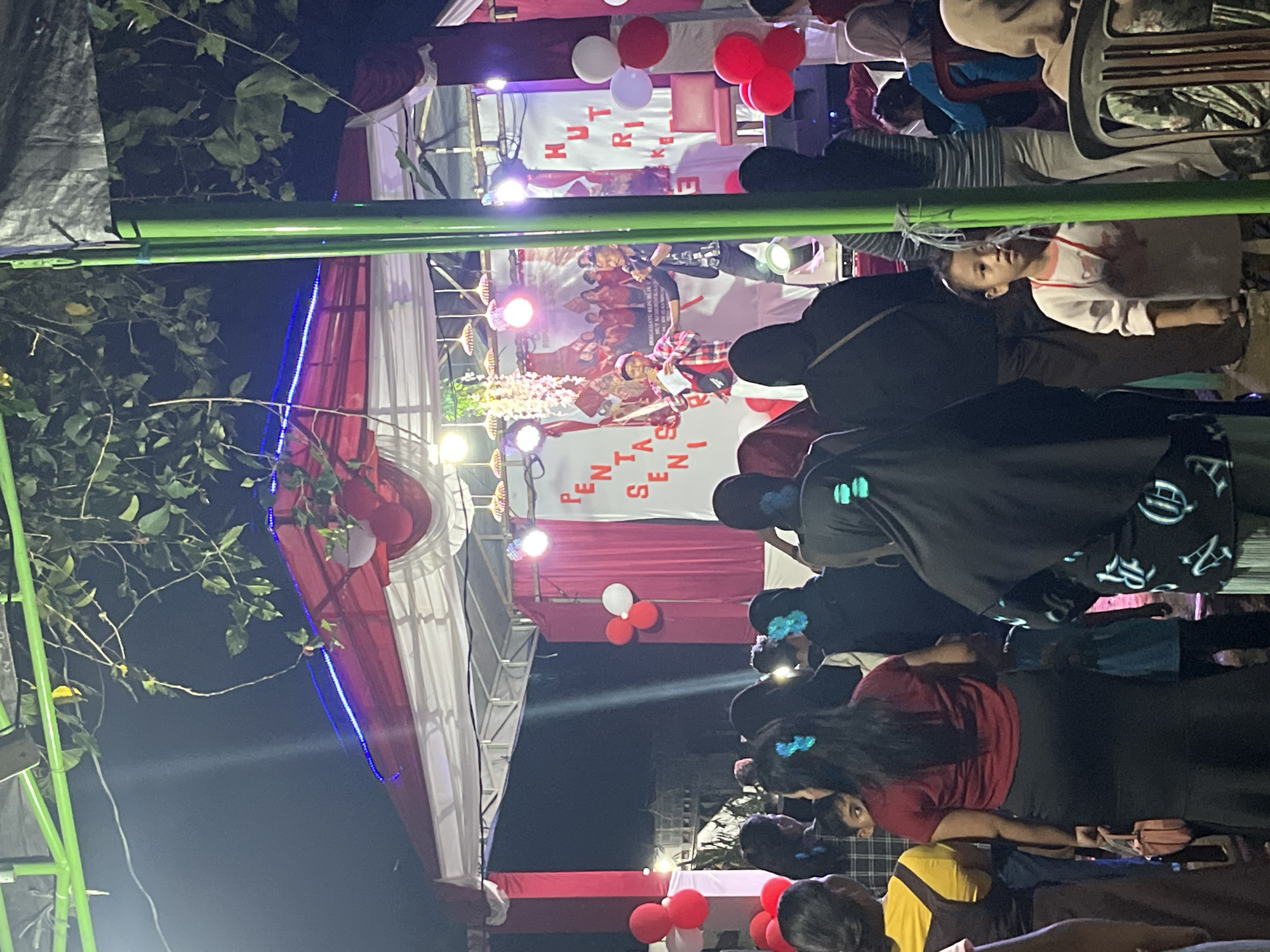 The image depicts an outdoor evening event in Tajurhalang, Indonesia, likely a community gathering or celebration, possibly related to a local anniversary given the Indonesian text visible on banners. A stage is set up, adorned with red and white drapes and illuminated by spotlights and blue LED lights. Two men are on the stage, one holding a microphone and speaking, the other holding papers and also appearing to speak. A crowd of people, including adults and children, are gathered in front of the stage, some standing and some seated on chairs. The foreground is dominated by the backs of several individuals, with a young child in a white shirt standing in the center of the frame, looking towards the stage. Balloons, in red and white, are visible as decorations around the stage. The surrounding environment appears to be a somewhat informal outdoor setting, possibly a field or open area, with some trees and greenery visible above the stage structure. The lighting suggests it is nighttime.
