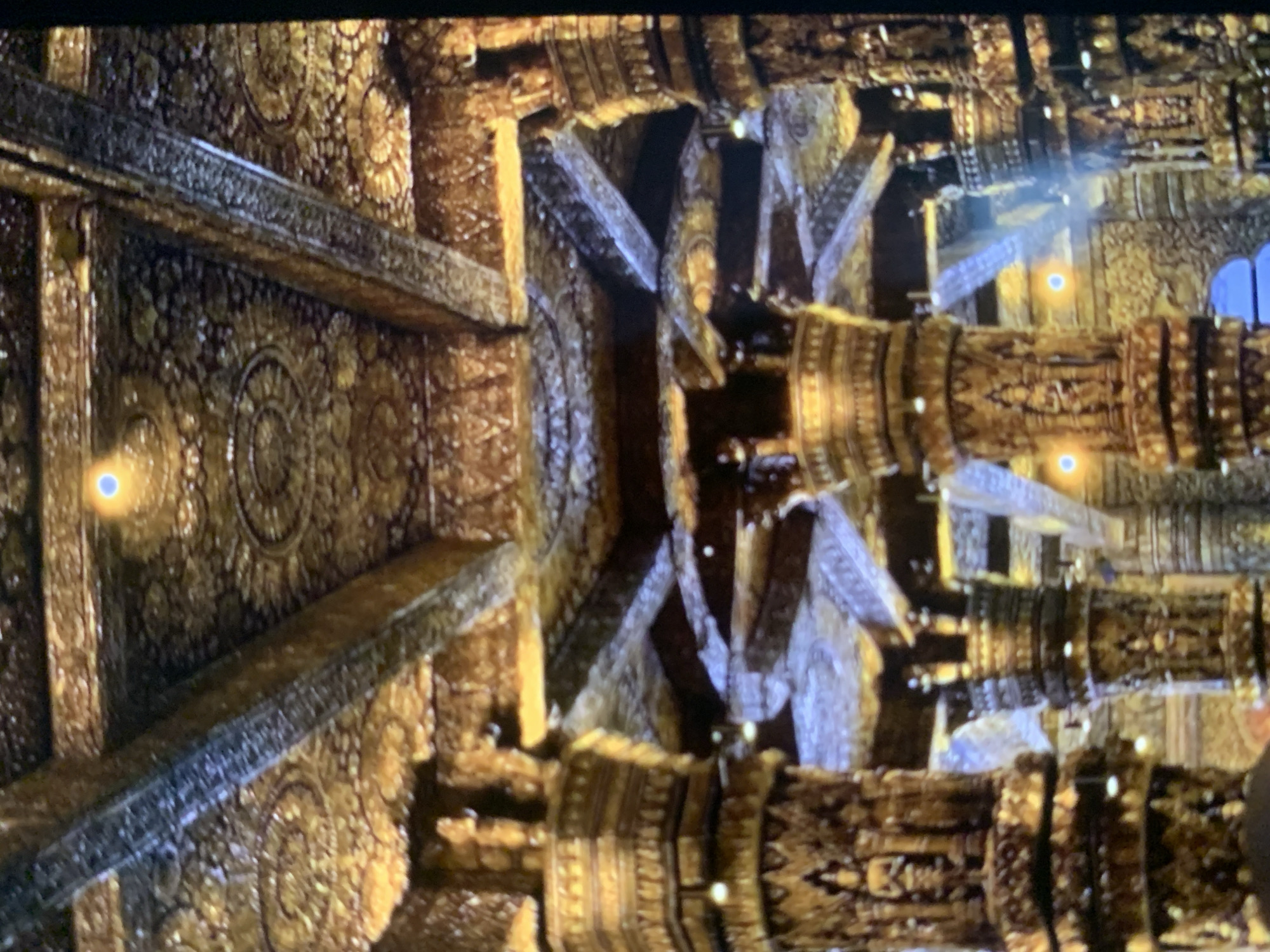 The image shows the interior of a richly decorated building, likely a temple or palace, with ornate golden pillars and a highly detailed ceiling. The pillars are carved with intricate patterns and figures, and the ceiling is adorned with what appear to be elaborate reliefs and possibly gold leaf. Lighting in the scene appears to be artificial, with some glowing orbs visible. The perspective suggests a wide-angle view looking upwards into the structure. There are no people or animals visible in the image. This scene is not located in Stavropol, Russia.