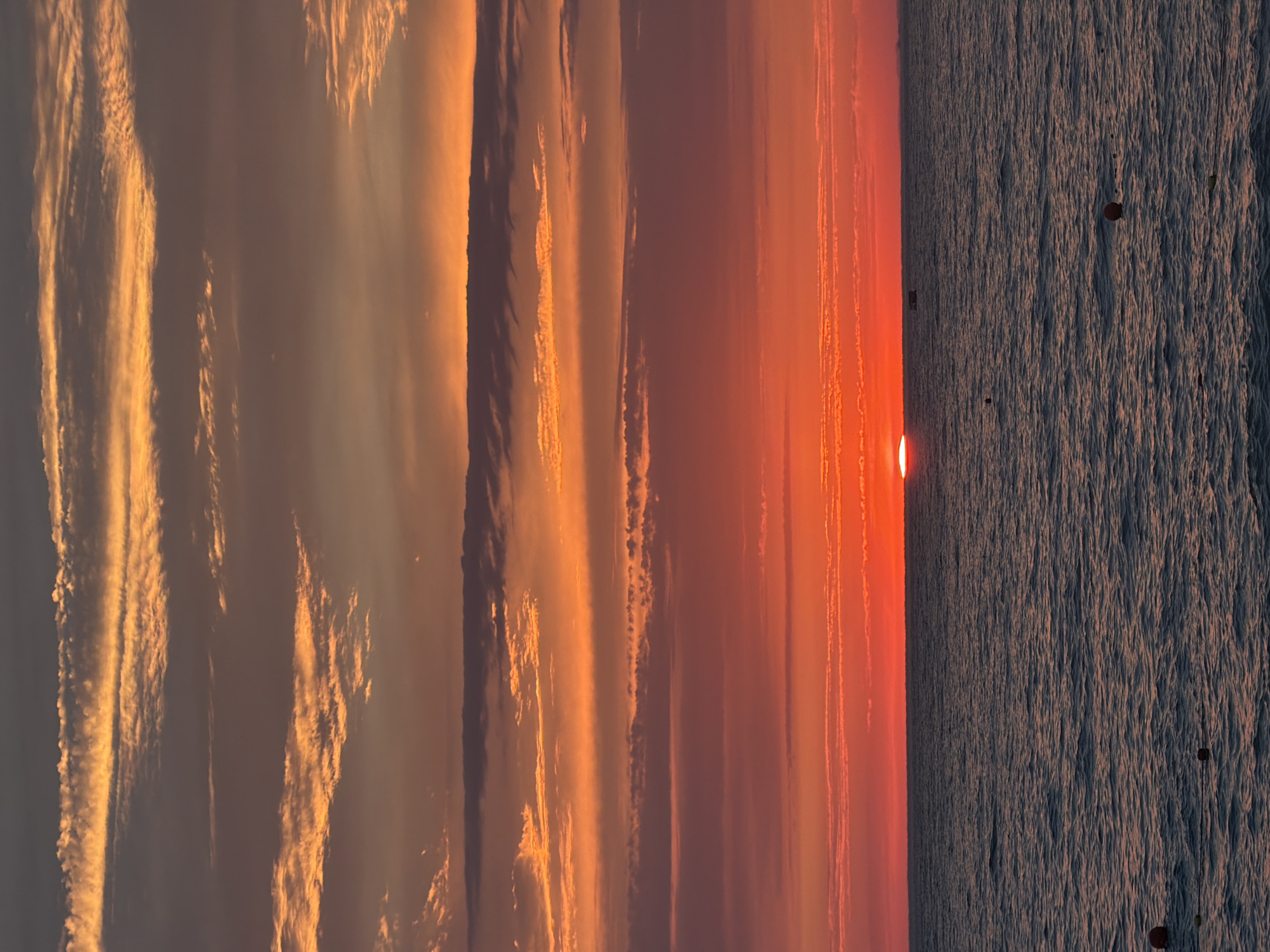 The image depicts a sunset over a body of water, likely the Black Sea, with Sochi, Russia, as the location context. The sun, a fiery orange orb, is just touching the horizon, casting a warm glow across the sky and water. The sky is a canvas of vibrant colors, with streaks of orange, pink, and gold from the setting sun blending with the soft blues and grays of the approaching twilight. Wispy clouds are illuminated by the sunlight, adding texture and depth to the atmospheric scene.
The water in the foreground is choppy, with visible ripples and waves creating a textured surface. Small, dark objects, possibly buoys or distant boats, are scattered across the water, adding a sense of scale and indicating it is an open expanse. The overall mood is serene and peaceful, evoking the tranquility of a sunset. The time of day is clearly evening, as indicated by the low position of the sun and the warm, diffused light. There is no visible text in the image.