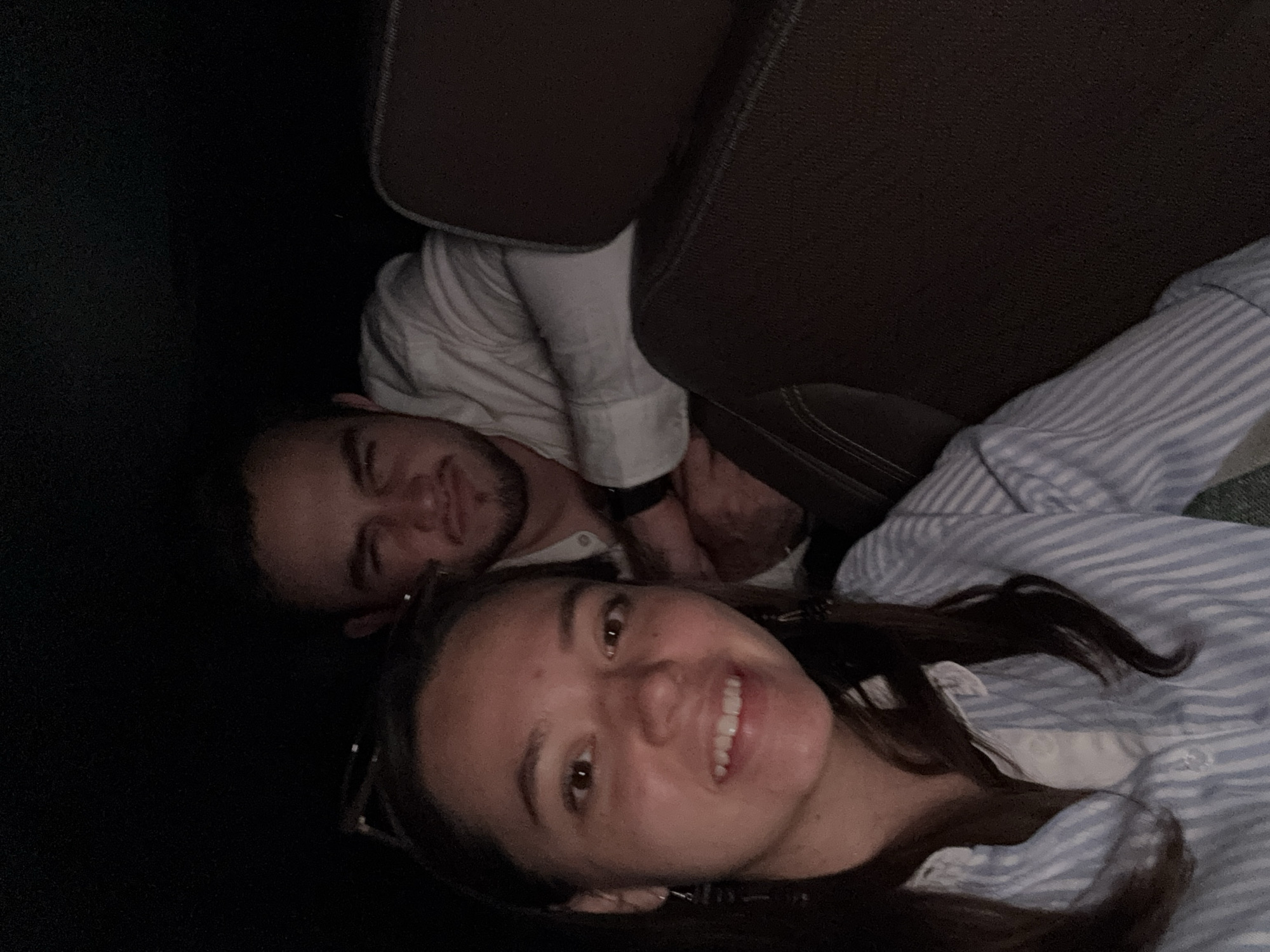 A diagonal, low-light photograph captures two individuals in what appears to be the interior of a vehicle or a dimly lit room, possibly in San Marcos Huixtoco, Mexico. In the foreground, a woman with dark hair and a bright smile looks directly at the camera. She has brown eyes and is wearing a light-colored, likely blue and white striped button-up shirt, with glasses perched on her head. Behind and slightly to her left, a man with a beard and dark hair is visible, looking towards the camera with a neutral expression. He is wearing a white long-sleeved collared shirt and a dark watch on his left wrist.
The setting is characterized by dark brown upholstery, possibly seat backs or headrests, framing the subjects. The background on the far left is deep black, indicating very low light conditions, suggesting it might be nighttime or a very dark interior space. The overall mood is casual and relaxed. No text is visible in the image.