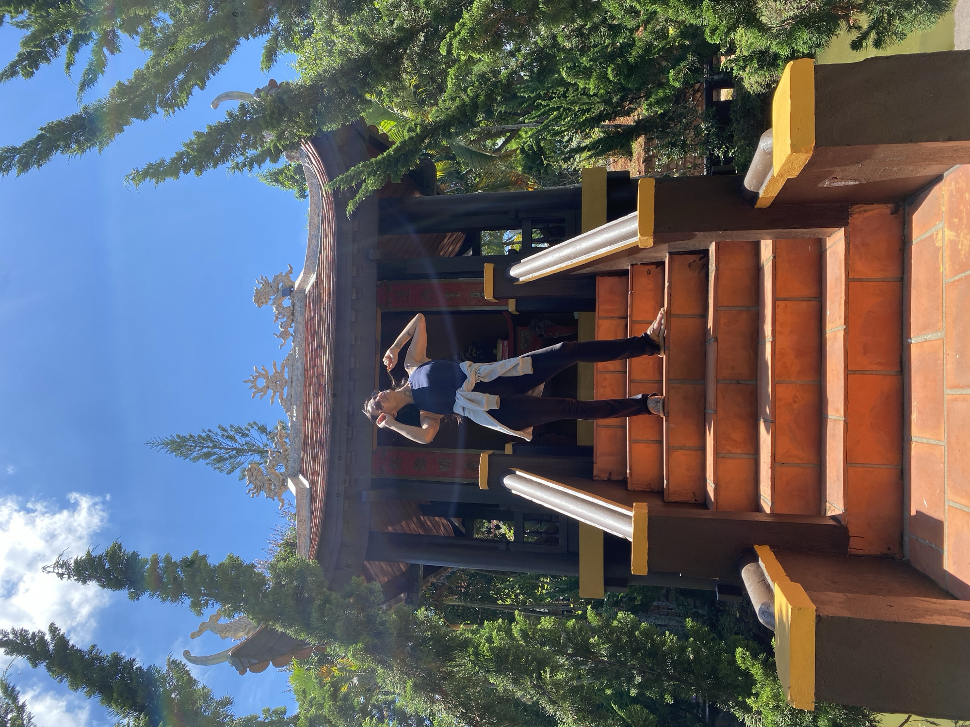 A person is performing yoga on a staircase in front of a temple. The person is wearing a blue tank top and dark pants, with a light-colored jacket tied around their waist. They are in a handstand pose, with their legs extended upwards and their arms supporting their body. The staircase is made of orange tiles and is bordered by a dark brown railing with yellow accents. The temple behind them has a tiled roof with intricate decorative carvings. Green trees and a bright blue sky with scattered clouds fill the background. The lighting suggests it is daytime, with the sun casting shadows on the stairs. The overall scene is serene and peaceful, likely in a tranquil outdoor setting in Vietnam, possibly Thôn 5. There is no visible text in the image.