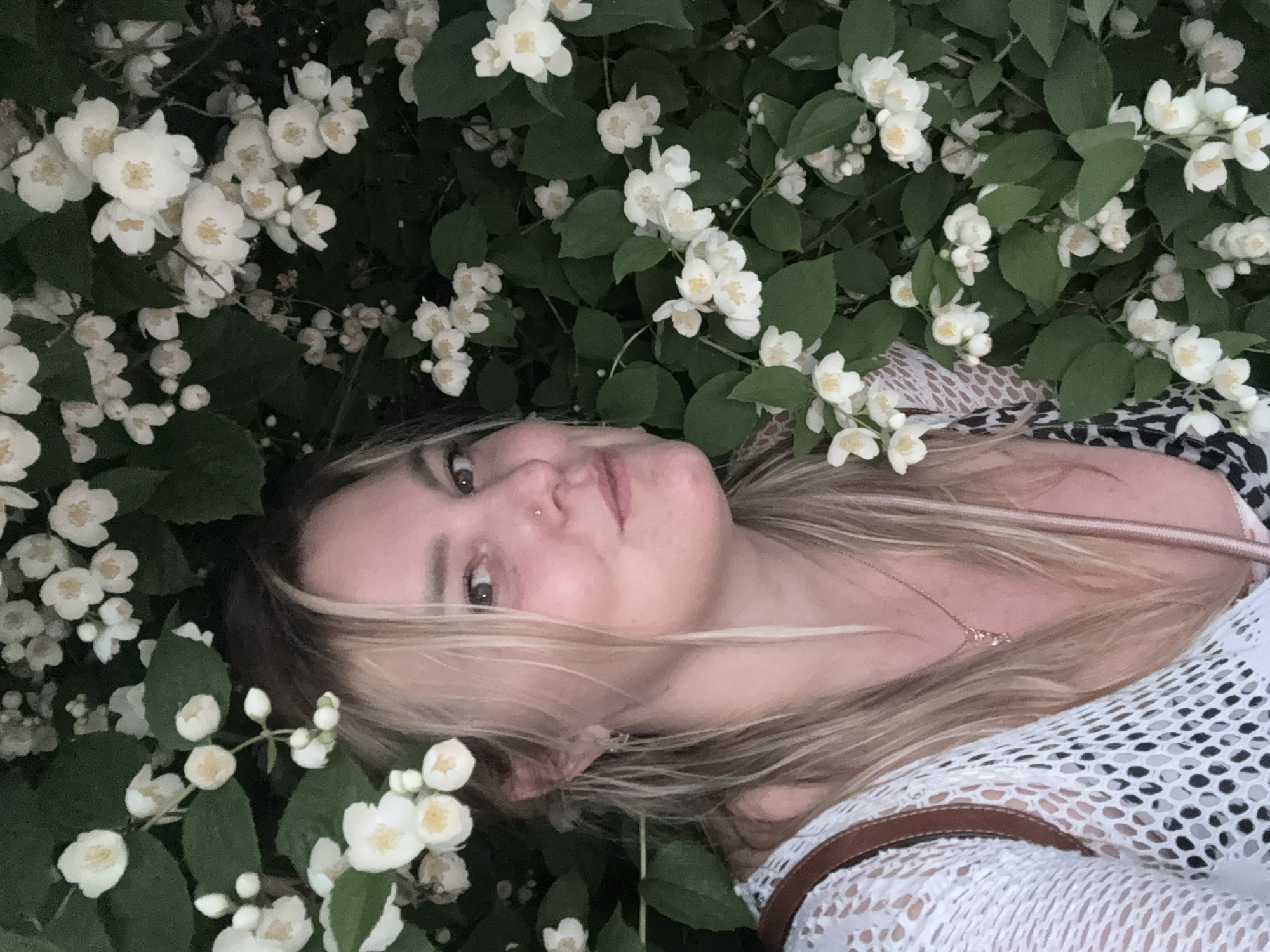 A blonde woman with fair skin and brown eyes is partially visible, nestled among abundant white jasmine flowers and green leaves. She is looking towards the camera with a slight smile. She is wearing a white crocheted top over a patterned dress and a thin necklace with a small pendant. The scene appears to be outdoors, possibly in a garden or park. There is no information available to determine the specific location within the city of Chaikovsky, Russia.