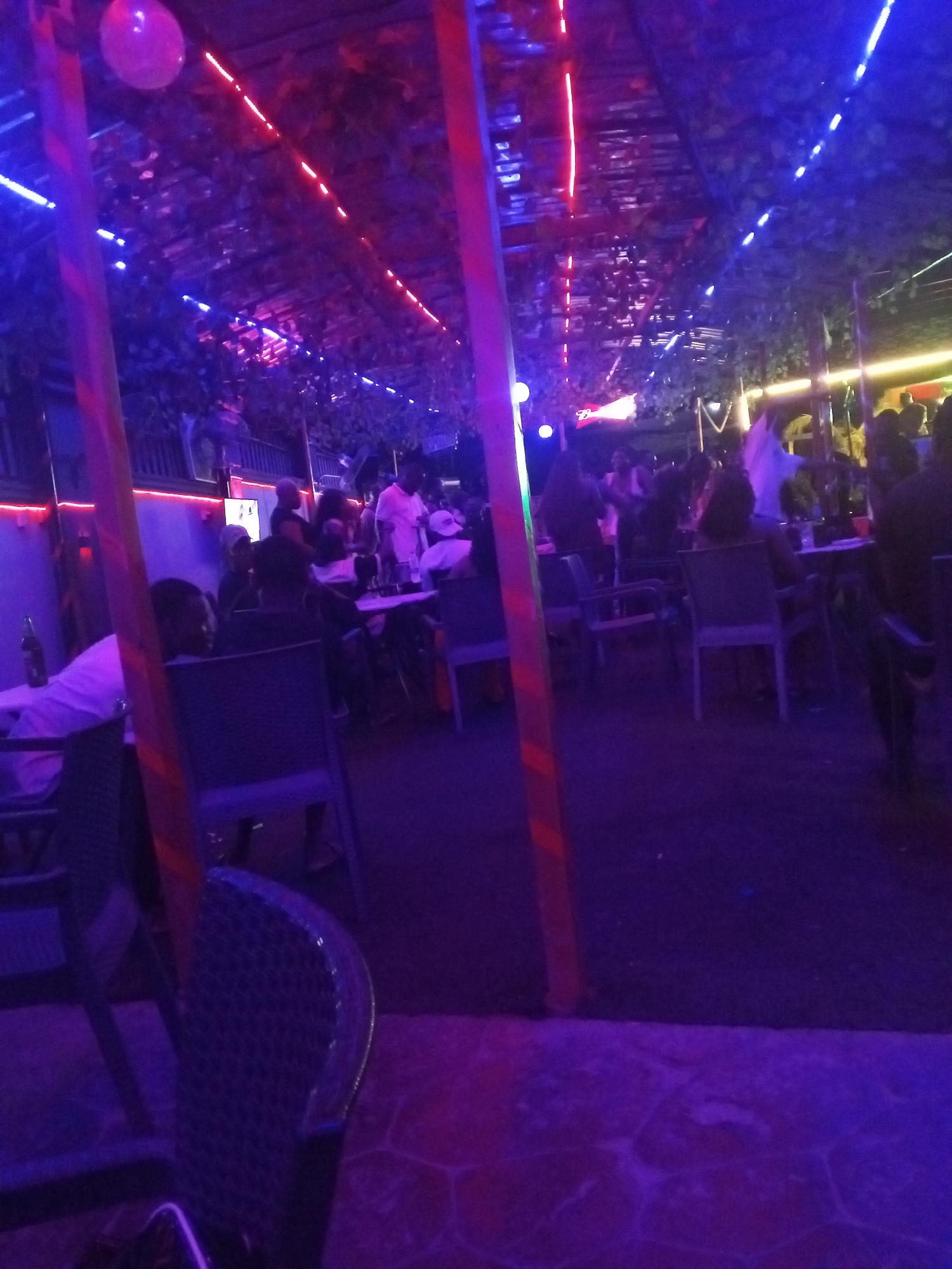 The image depicts an outdoor bar or restaurant scene at night in Lokoja, Nigeria. The space is illuminated with blue and red neon lights strung across the ceiling, which is covered with vines or artificial foliage. Several people are seated at tables, and others are standing or walking around. The chairs appear to be made of plastic or wicker. The floor is patterned with a tile or stone design. The atmosphere is lively and social.