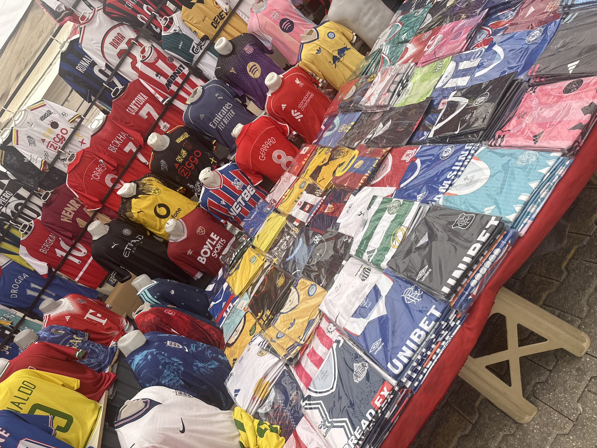 The image shows a street vendor selling a variety of replica soccer jerseys in Fethiye, Turkey. The jerseys are displayed on a table and hanging from a metal rack. The jerseys represent various teams and players, including famous names like Ronaldo, Beckham, and Henry. Some jerseys are on small mannequin torsos, while others are individually wrapped in plastic. The table is covered with a red cloth. The stand is supported by a folding plastic table leg. The ground is paved with gray tiles.