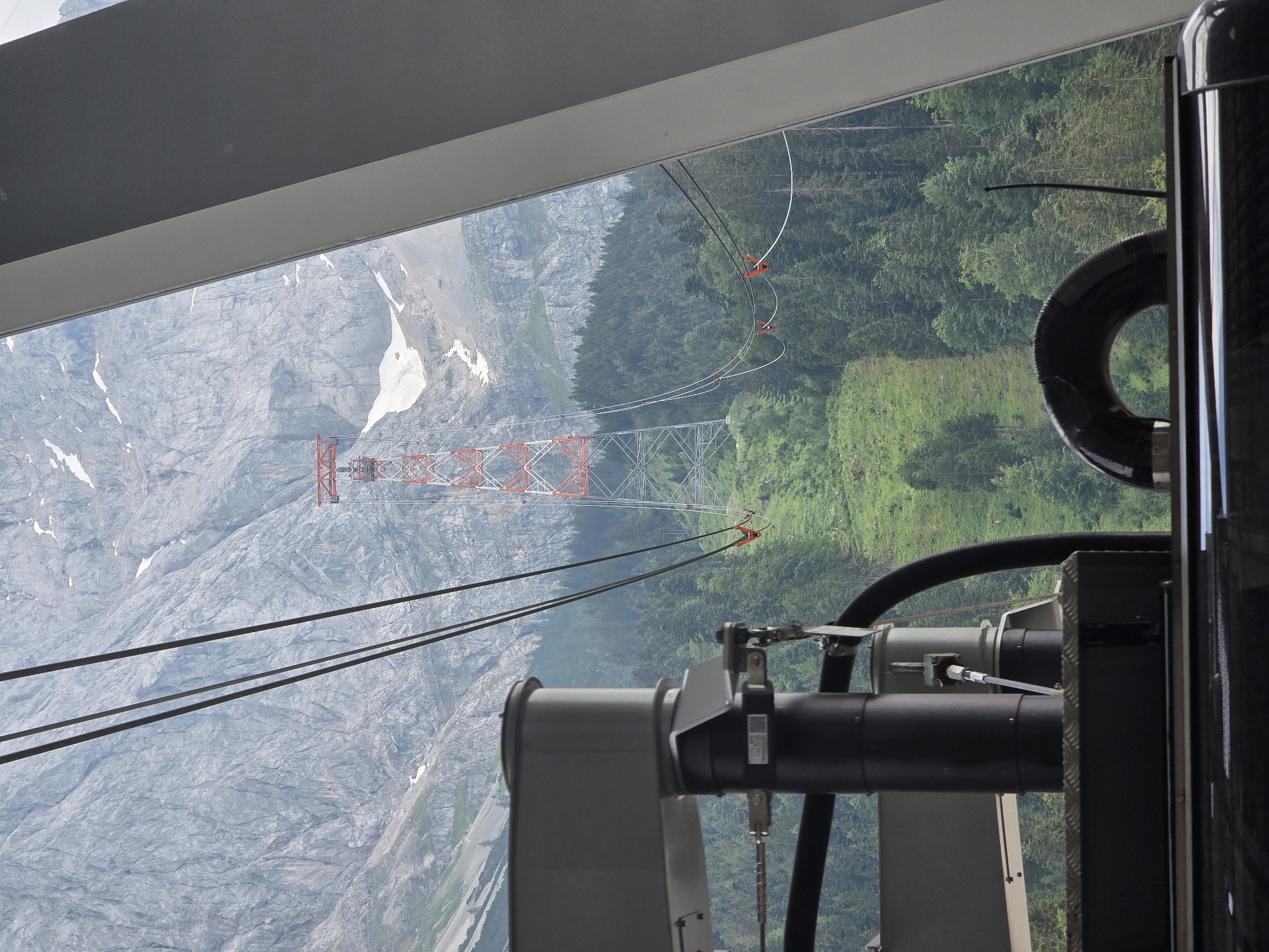 This image captures a view from a cable car station in Grainau, Germany, looking out towards the Zugspitze mountain range. The foreground is dominated by the mechanical components of the cable car system, including dark pipes, metal structures, and cables. In the midground, a vast expanse of evergreen forest covers the slopes of the mountains, with patches of green grass on a steep incline. A prominent red and white lattice tower, a support for the cable car, stands tall against the backdrop of the rugged, grey mountain peaks, some of which are still dusted with snow. The sky is overcast, casting a soft light on the scene. The perspective suggests the viewer is looking out from a modern, enclosed structure.
