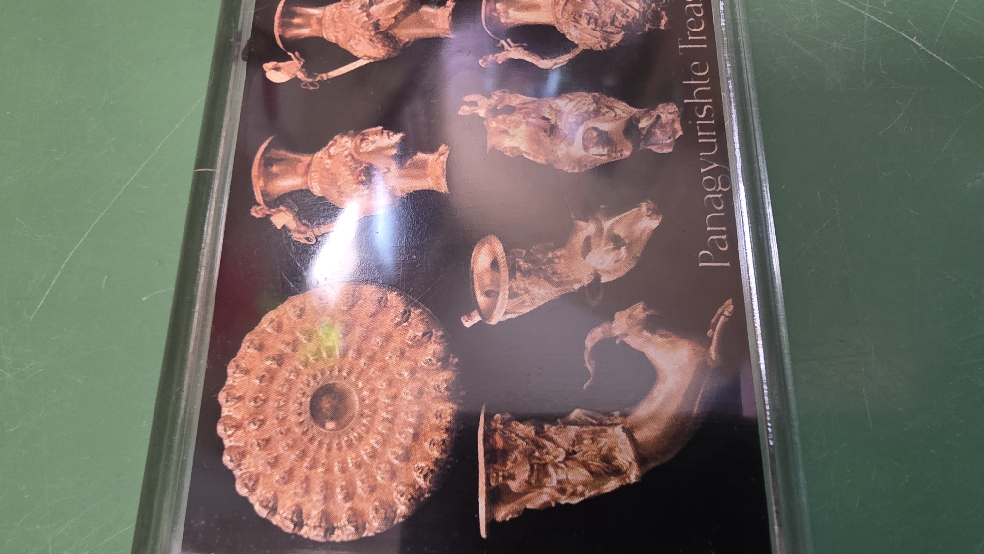 A photograph depicts an encased image of artifacts, labeled "Panagyurishte Treasure," suggestive of a museum or display of Bulgarian cultural heritage. The artifacts are of a golden or bronze hue, arranged against a dark backdrop. The image includes animal-shaped drinking horns (rhytons) and other ornamental objects. The casing around the image appears to be made of clear plastic or glass, possibly to protect the display. The background is a solid green color, likely a table or other surface the encased image rests upon. Location in the city of Blagoevgrad, Bulgaria, cannot be determined from this image.