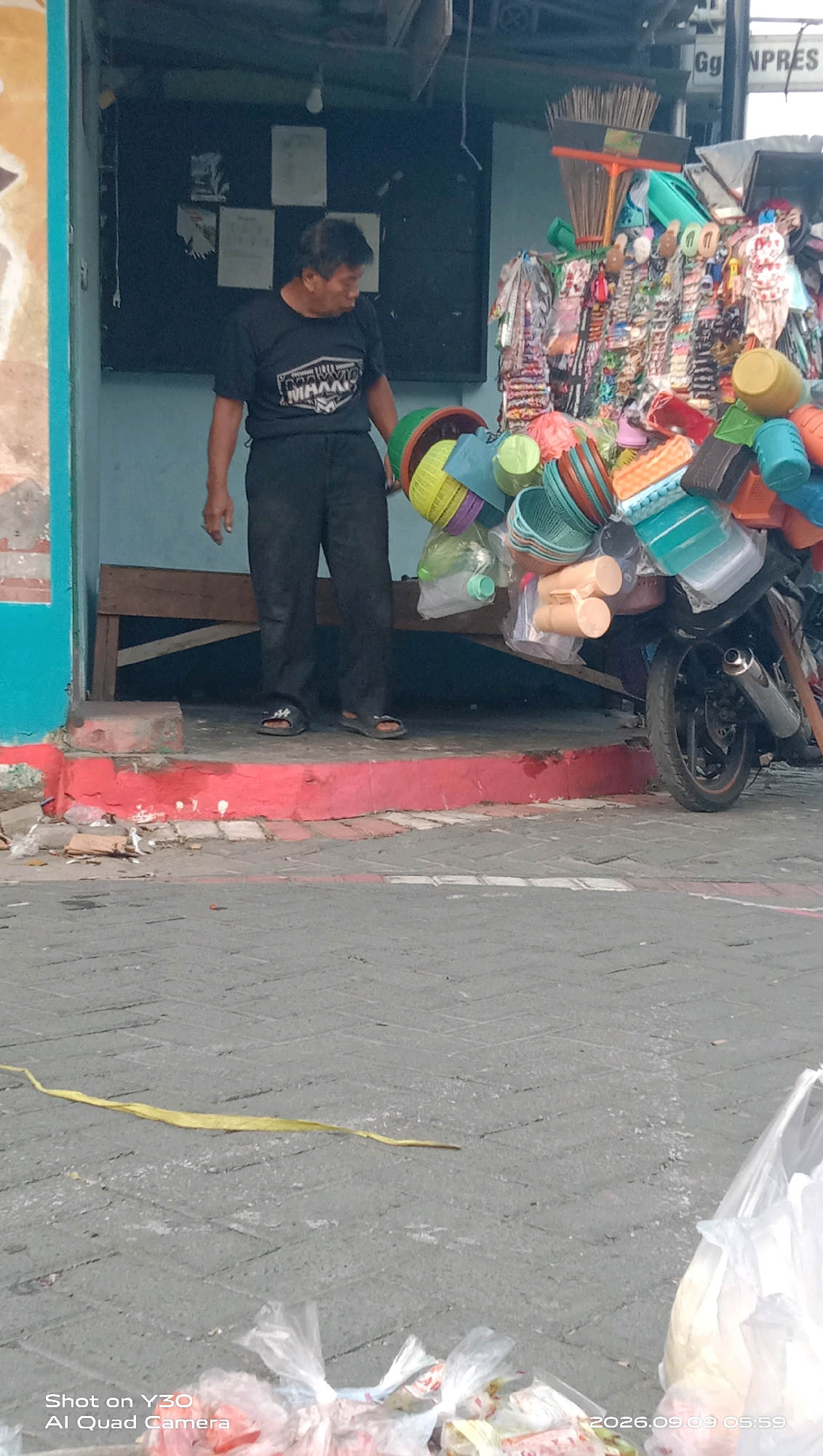 In Klangonan, Indonesia, a man in black t-shirt and pants stands near a makeshift stall constructed from a motorcycle loaded with household items. The stall is adorned with an array of colorful plastic containers, buckets, and brooms, suggesting a small business operation. The man appears to be a vendor, possibly preparing to sell or having just finished selling his merchandise. The surrounding environment shows a tiled street and a simple building, hinting at a commercial or residential area within the city.