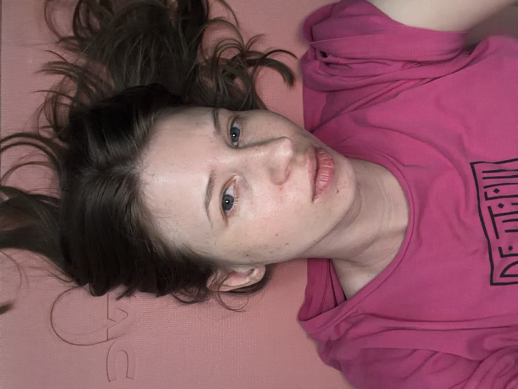 The image shows a woman lying on a pink yoga mat. She has long, dark brown hair spread out around her head. Her eyes are open and looking towards the camera, and her lips are slightly parted. She is wearing a bright pink t-shirt with a black graphic design on the front. The background is a solid pink surface, which appears to be the yoga mat. The lighting is soft and appears to be coming from above. There are no visible indications of the city of Dongducheon-si, South Korea, in the image.