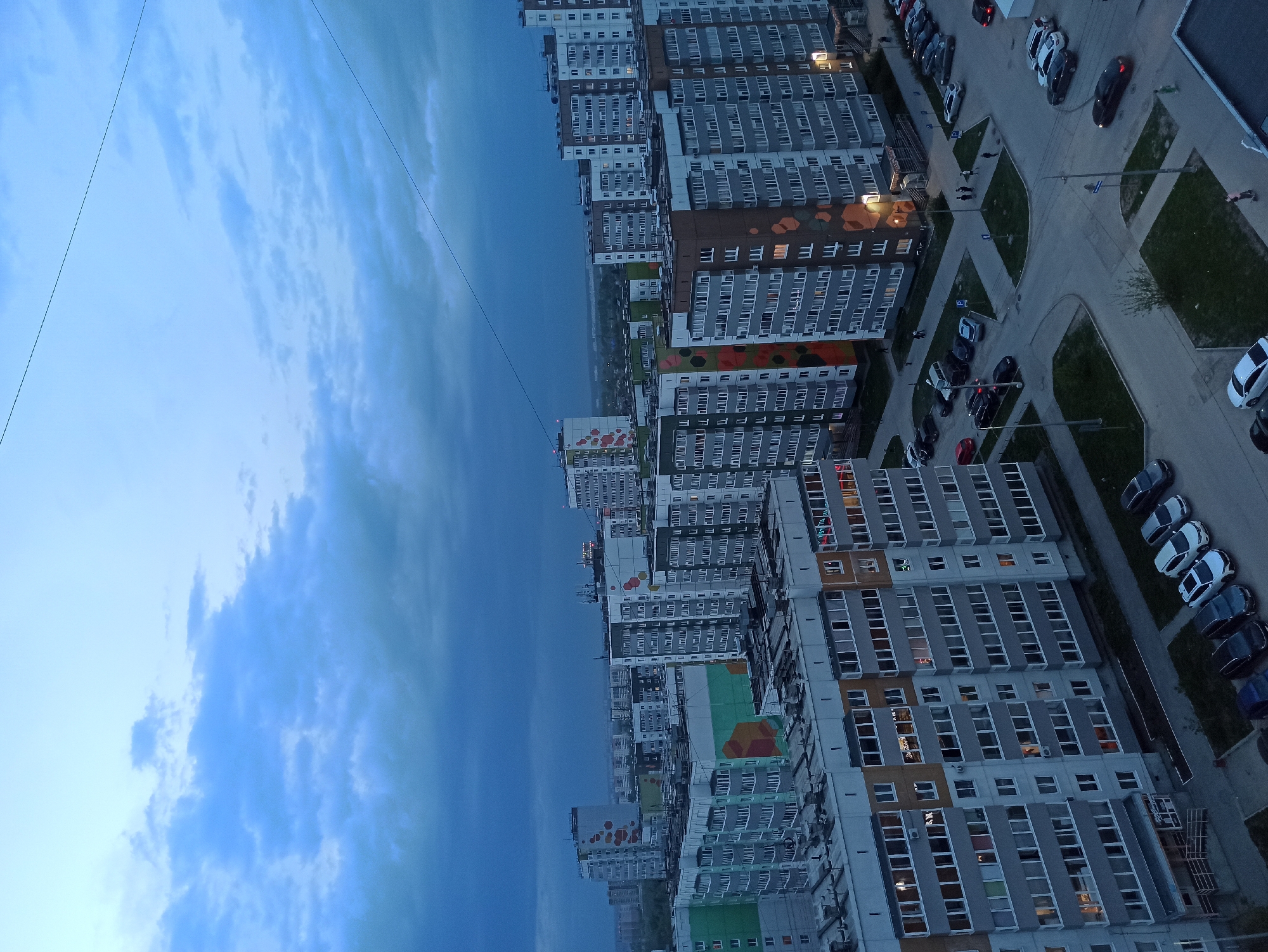 The image shows an aerial view of a residential area in Кондратово, Russia, at dusk. Multiple high-rise apartment buildings are visible, arranged in a row. The buildings have repetitive window patterns and some are adorned with colorful murals. Roads and parking areas are seen between the buildings, with several parked cars. The sky is overcast with a blue hue. Power lines are visible crossing the sky. The overall atmosphere is urban and residential.