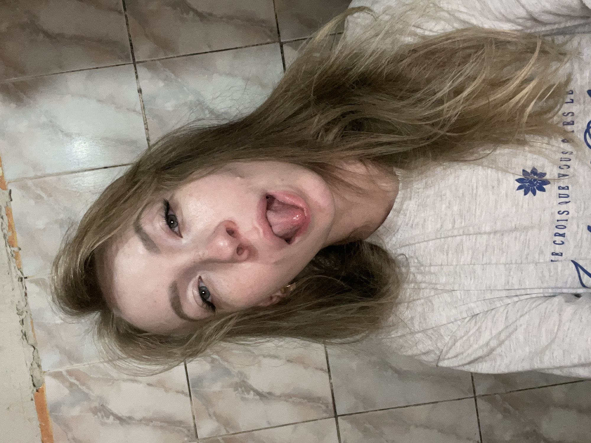 A young woman with fair skin and light brown hair is lying on a tiled floor. Her head is tilted to the left, and she is sticking her tongue out. She is wearing a light gray t-shirt with blue flower prints and some words written on it. The tiles are a light beige color with a marble-like pattern. Based on available information, it is not possible to confirm the location in the city of Perm, Russia.