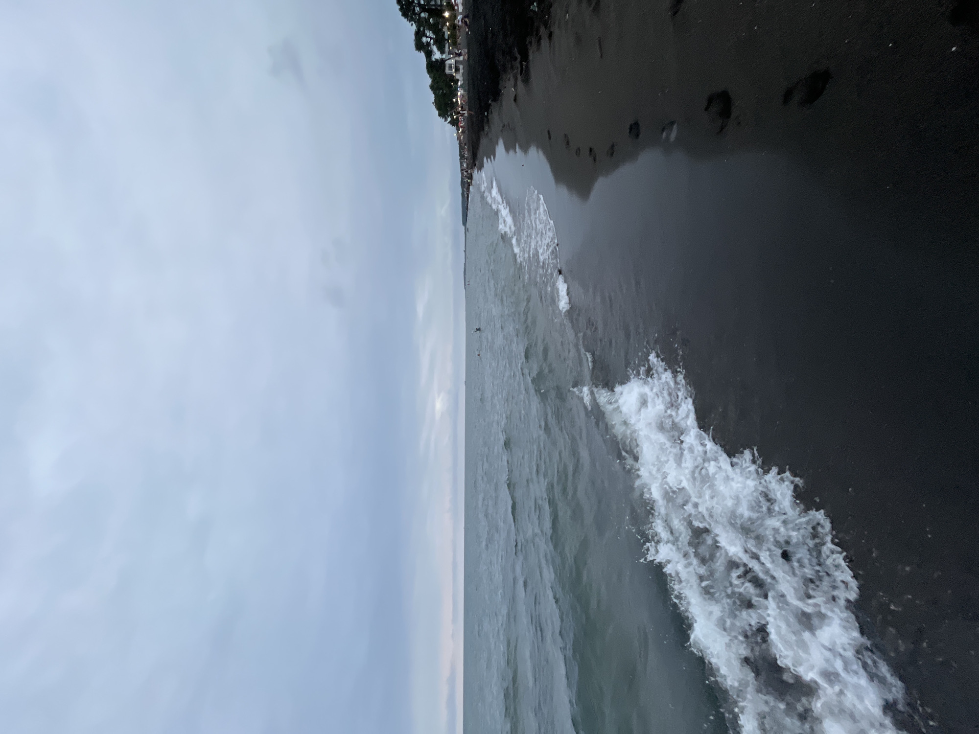 The image shows a view of the Ureki beach in Georgia. The sky is overcast with gray clouds. The sea is choppy with moderate waves. The beach is covered in dark sand, and there are footprints visible on the wet sand near the water's edge. In the distance, along the shoreline, there are trees and lights, suggesting a developed area with some people present. One person can be seen swimming in the water.