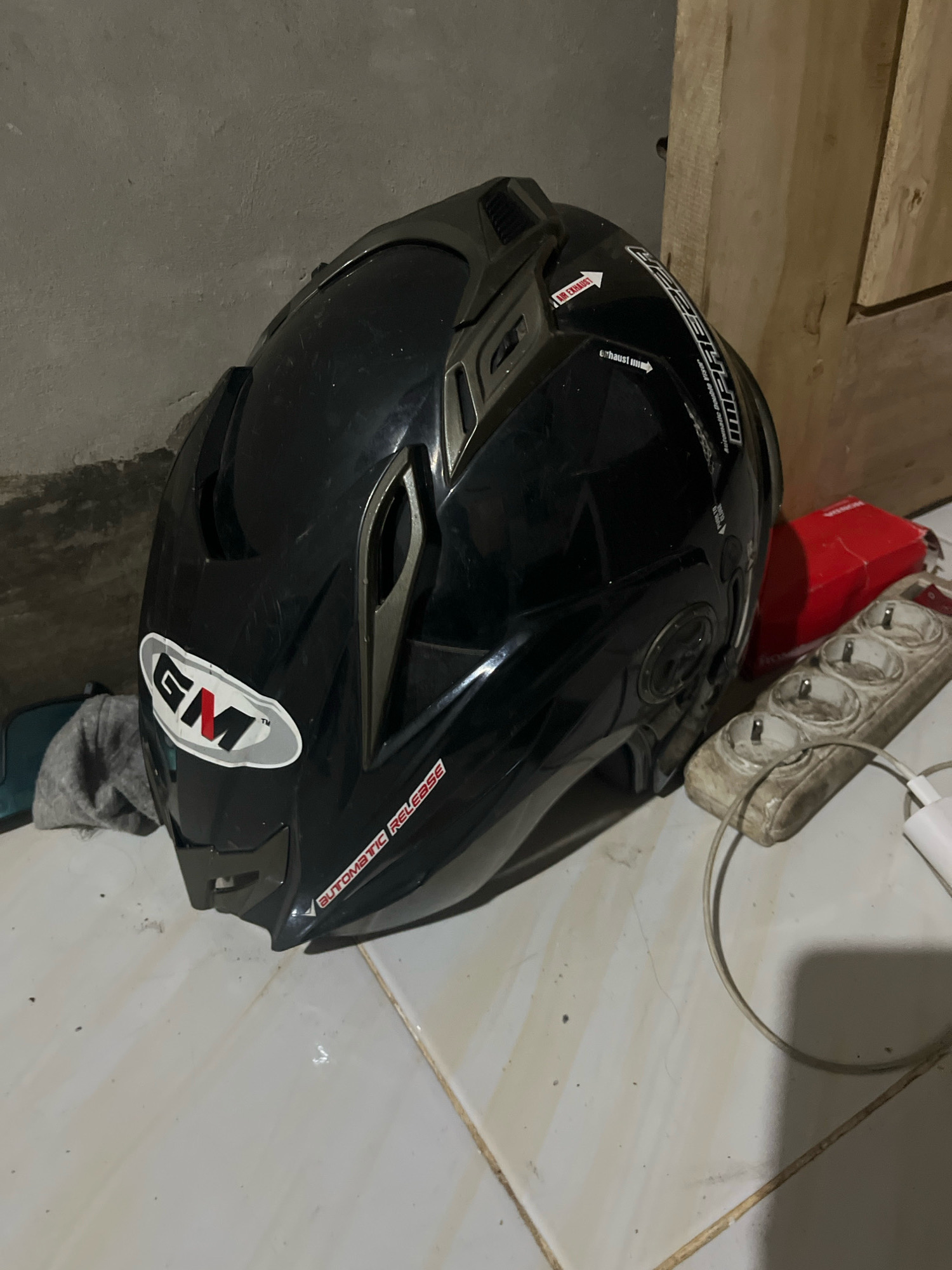 A black motorcycle helmet is lying on its side on a tiled floor in what appears to be an indoor space with a rough, grey wall and a wooden partition in the background. The helmet has several stickers, including a prominent oval "GM" logo with red and white lettering, and other decals with text such as "AIR EXHAUST" and "AUTOMATIC RELEASE." A multi-outlet power strip with several plugs and a white charging cable are visible to the right of the helmet. A red box is also present near the power strip. The floor is tiled and appears somewhat dirty, with scattered dust or debris. The lighting is dim, suggesting an indoor environment possibly in Kebumen, Indonesia, given the context provided.