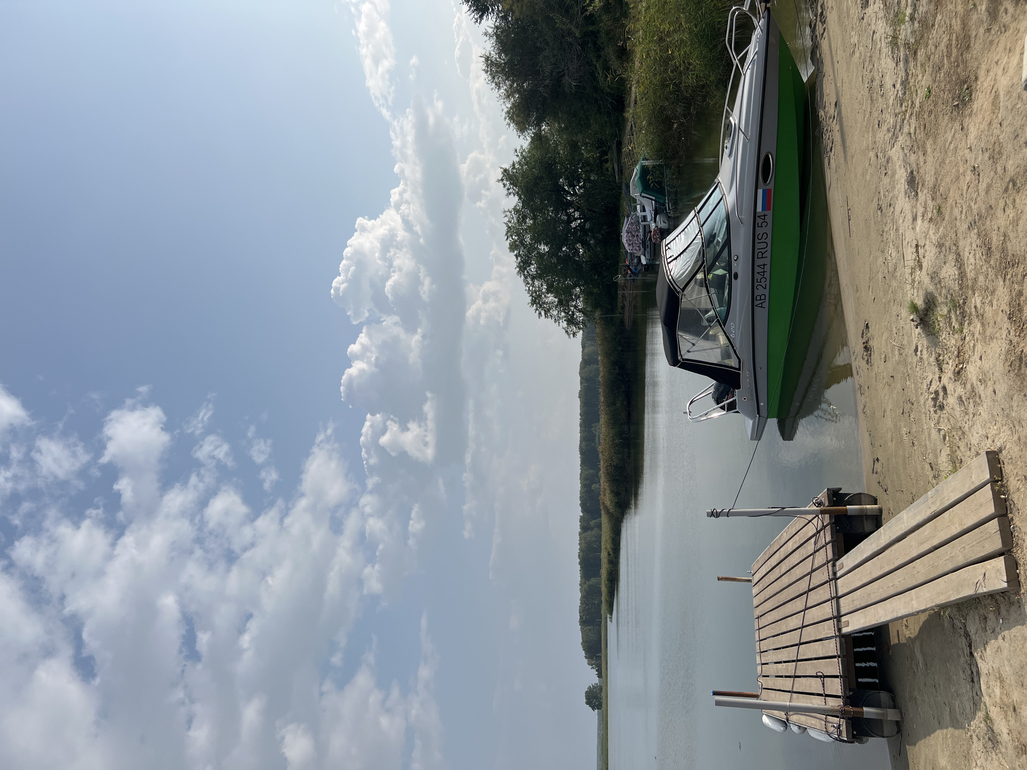 A motorboat with the registration number "AB 2544 RUS 54" is docked on a sandy shore next to a wooden pier on a calm body of water. Tall, dry reeds line the opposite bank, beyond which a dense forest of green trees stretches towards the horizon. The sky above is a pale blue, dotted with scattered white clouds. The scene suggests a serene, natural setting likely in the vicinity of Novopichugovo, Russia, a rural settlement known for its natural landscapes and waterways. There are no people visible in the image, and the focus is on the boat, the pier, and the natural environment. The water appears still, reflecting the sky and surrounding vegetation. The overall atmosphere is peaceful and tranquil.